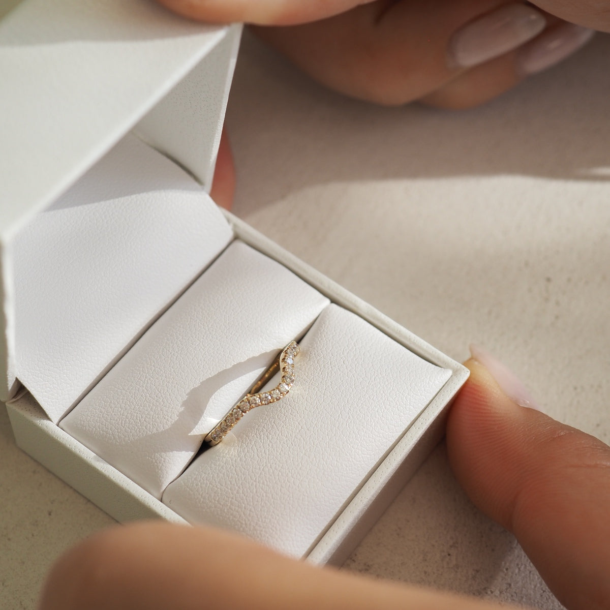 Elegant gold diamond ring in white jewellery box, held by hands with manicured nails, placed on a soft, light surface.