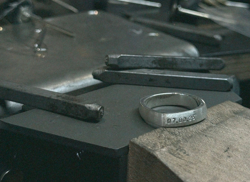 A silver metal band ring engraved with the date '07.02.95' resting on a dark anvil in a workshop setting.