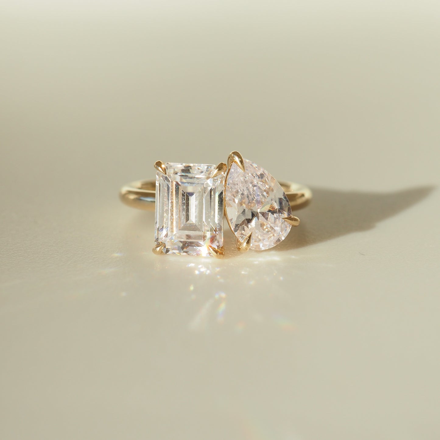 Gold ring featuring an emerald-cut and pear-shaped diamond, elegantly set side by side, casting a soft shadow on a smooth surface.