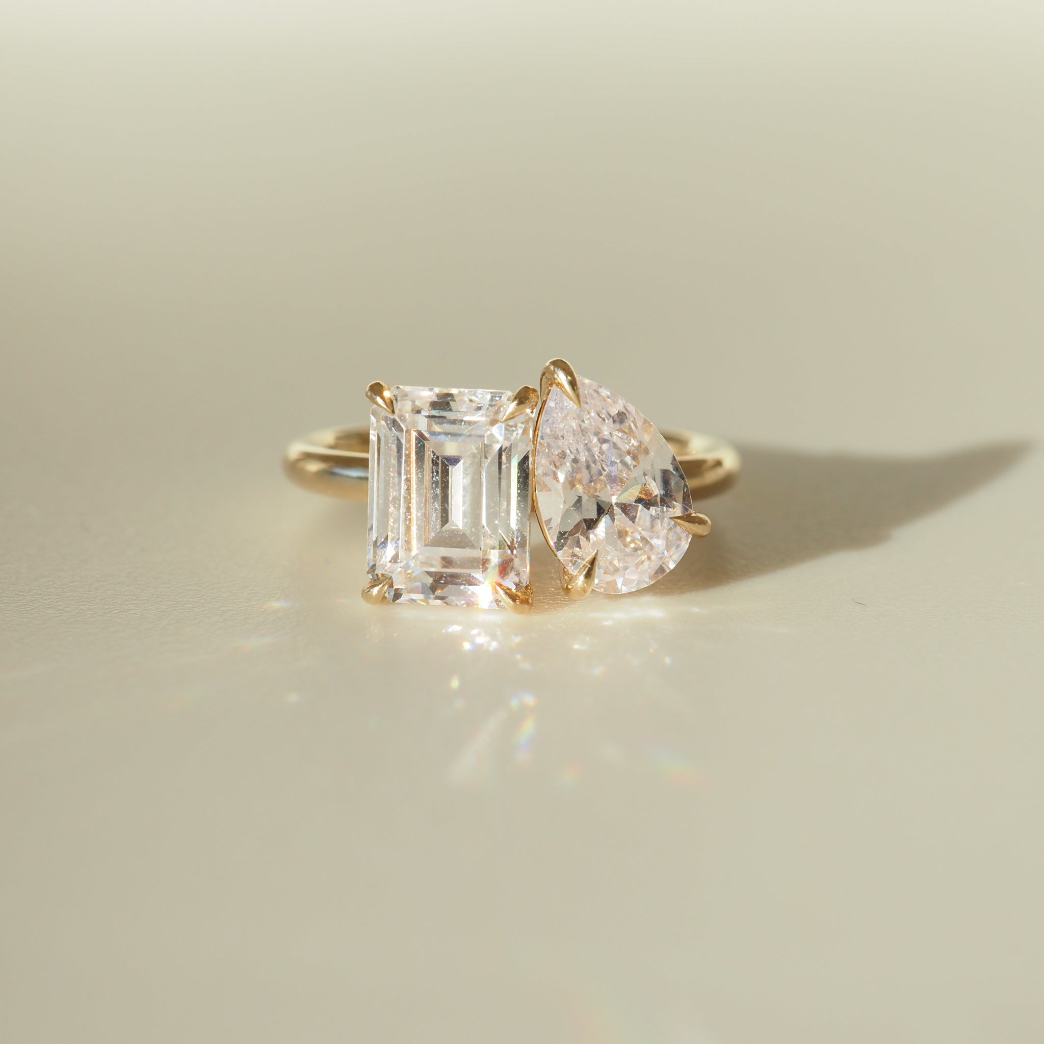 Gold ring featuring an emerald-cut and pear-shaped diamond, elegantly set side by side, casting a soft shadow on a smooth surface.