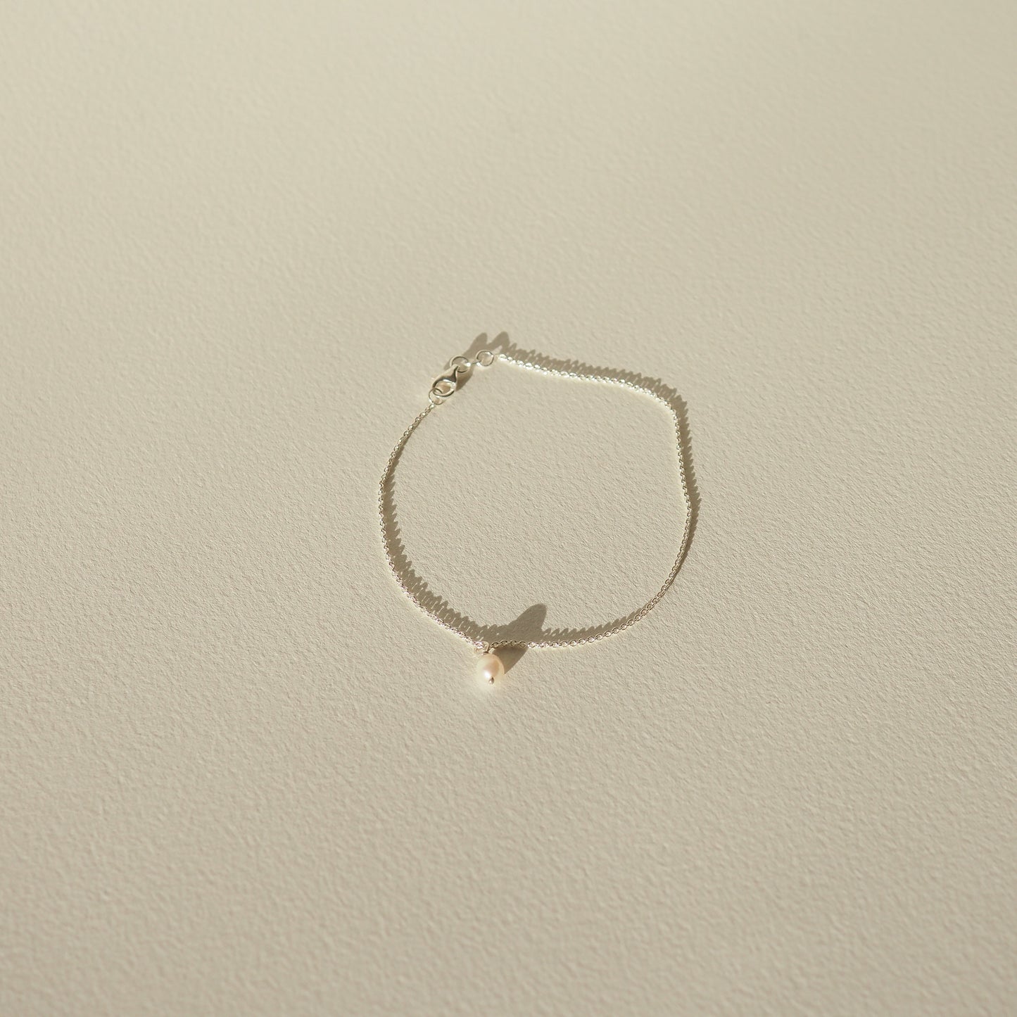 Delicate gold chain bracelet with a single pearl pendant, casting a soft shadow on a light-coloured surface. Minimalist luxury jewellery.