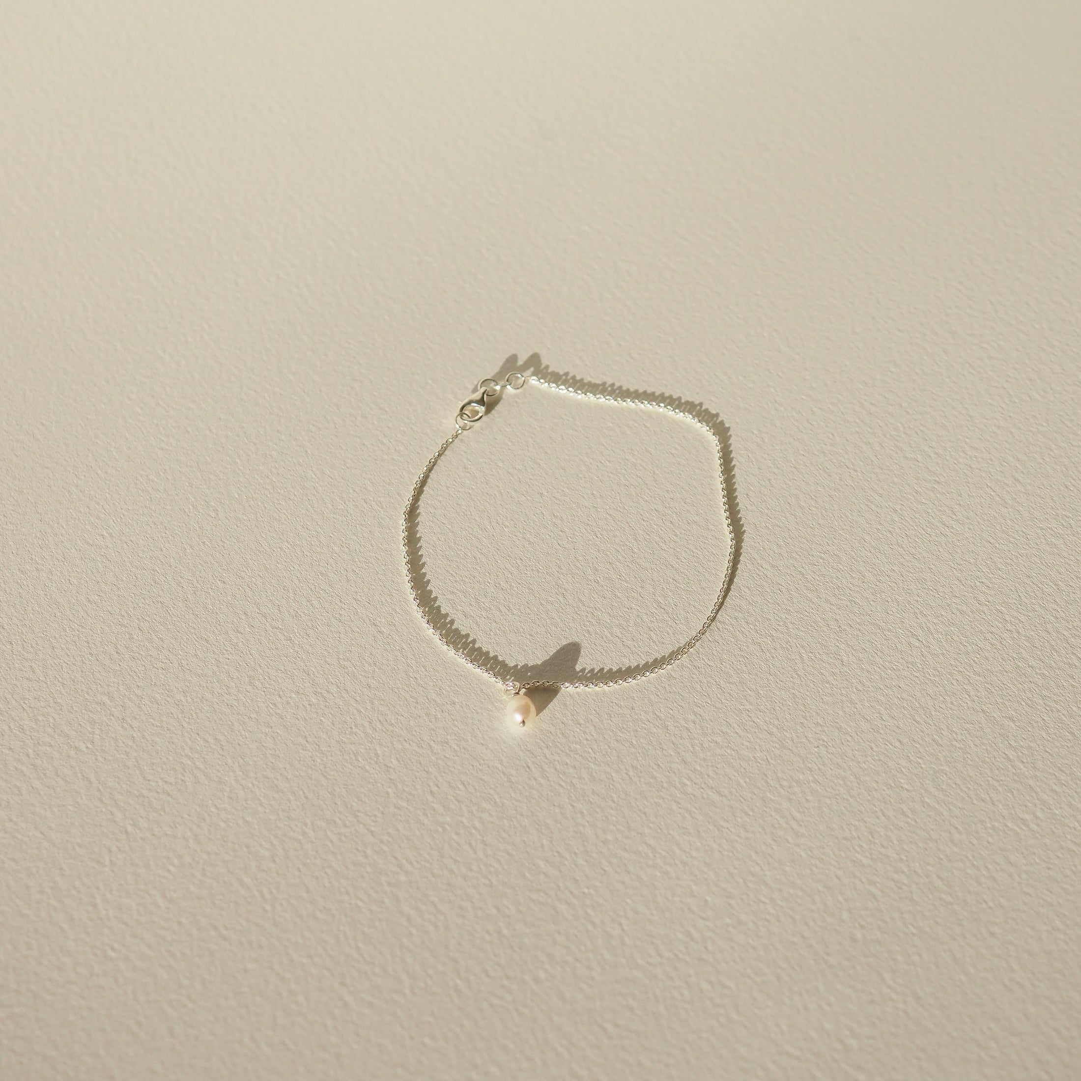 Delicate gold chain bracelet with a single pearl pendant, casting a soft shadow on a light-coloured surface. Minimalist luxury jewellery.