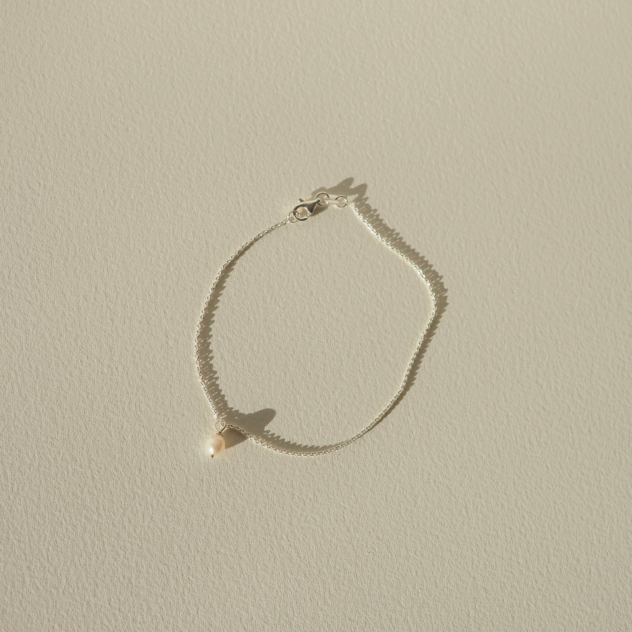 Delicate silver bracelet with a single pearl on a light textured background, casting a soft shadow. Perfect for luxury jewellery lovers.