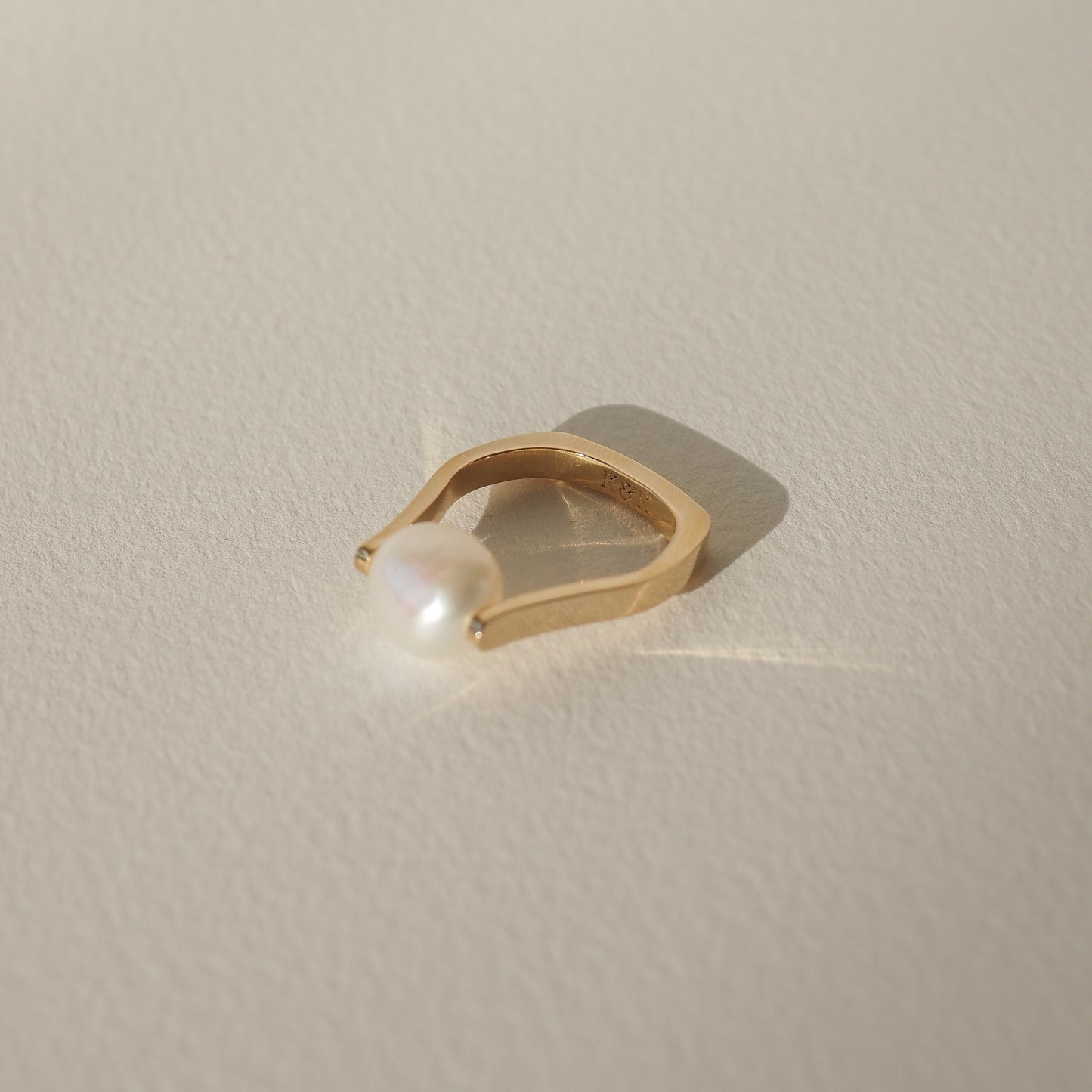 Gold ring with a minimalist design, featuring a single, luminous pearl. The ring casts a subtle shadow on a light, textured surface.