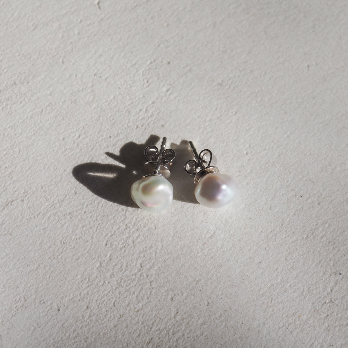 Two lustrous pearl stud earrings on a light textured surface, casting subtle shadows. Perfect for high-end jewellery enthusiasts.