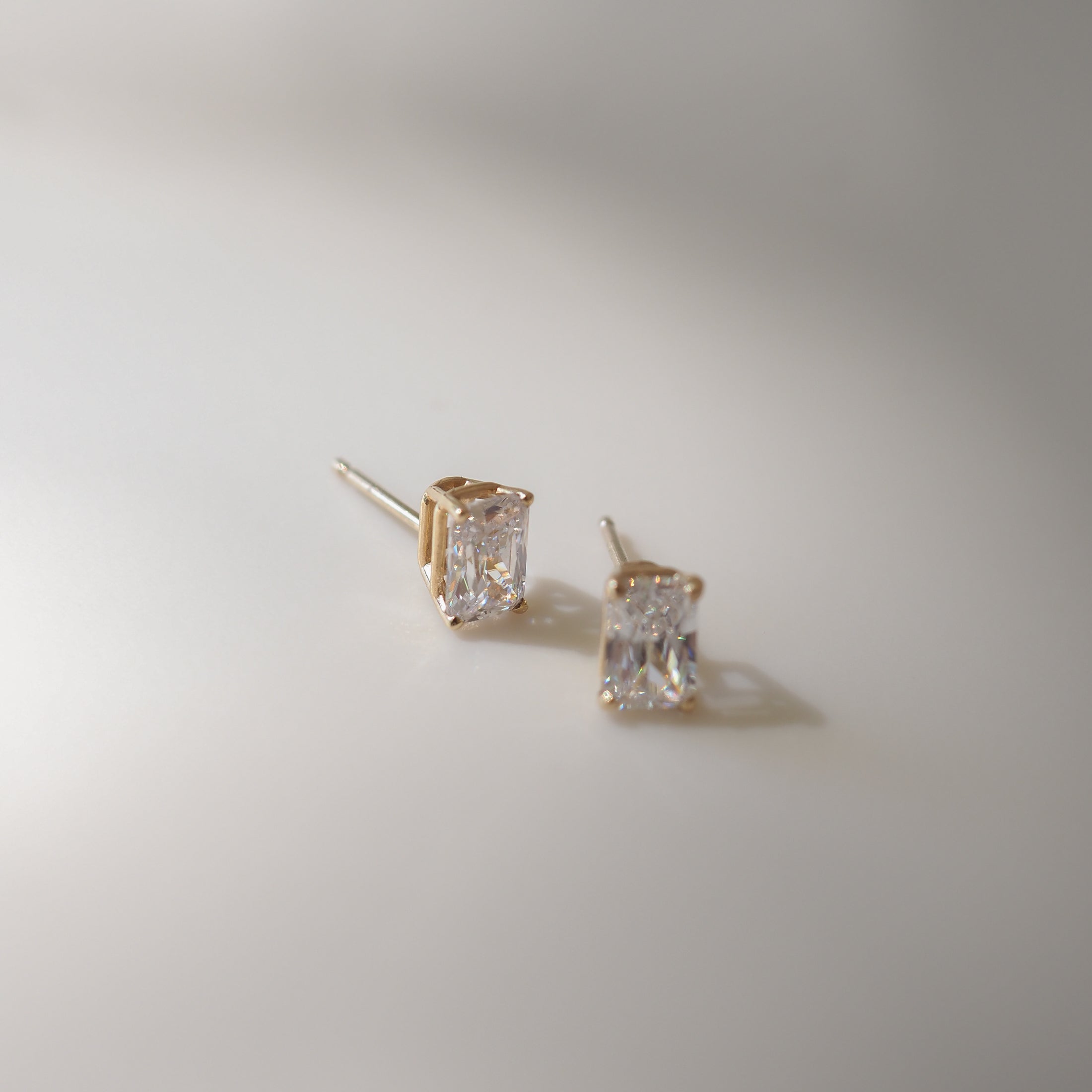 Two radiant-cut diamond stud earrings set in gold, elegantly displayed on a smooth, light-background surface.