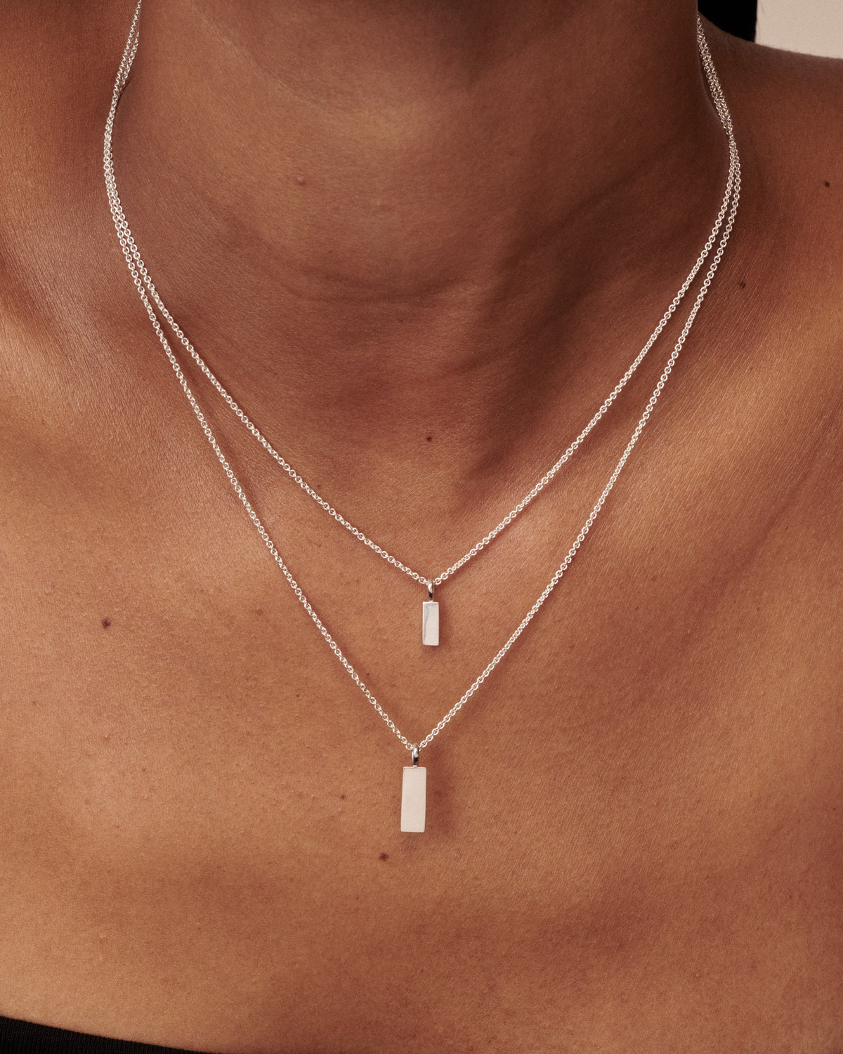 A close-up of a person's neck and upper chest wearing a delicate double-layered silver necklace with rectangular bar pendants.