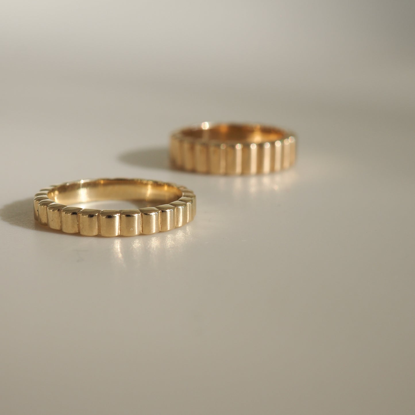 Two elegant gold rings with grooved patterns, placed on a smooth surface, casting soft shadows. Luxurious jewellery display.