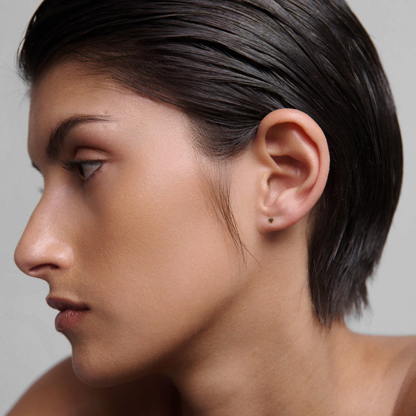Profile of a person with slicked-back hair wearing a subtle, small stud earring. Skin appears smooth and makeup is minimal.