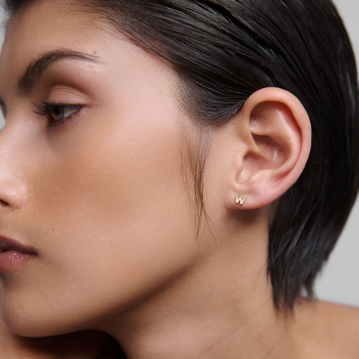 Close-up of a woman's ear wearing a small gold "W" initial earring; luxury jewellery, minimalist design, sleek style.