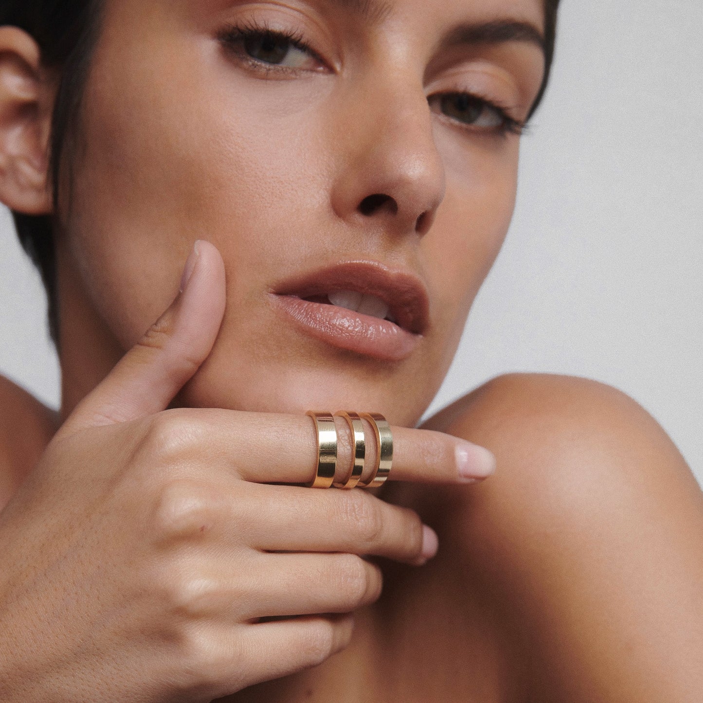 Woman's hand with luxurious, stacked gold rings held near her lips, showcasing elegant jewellery against a neutral background.
