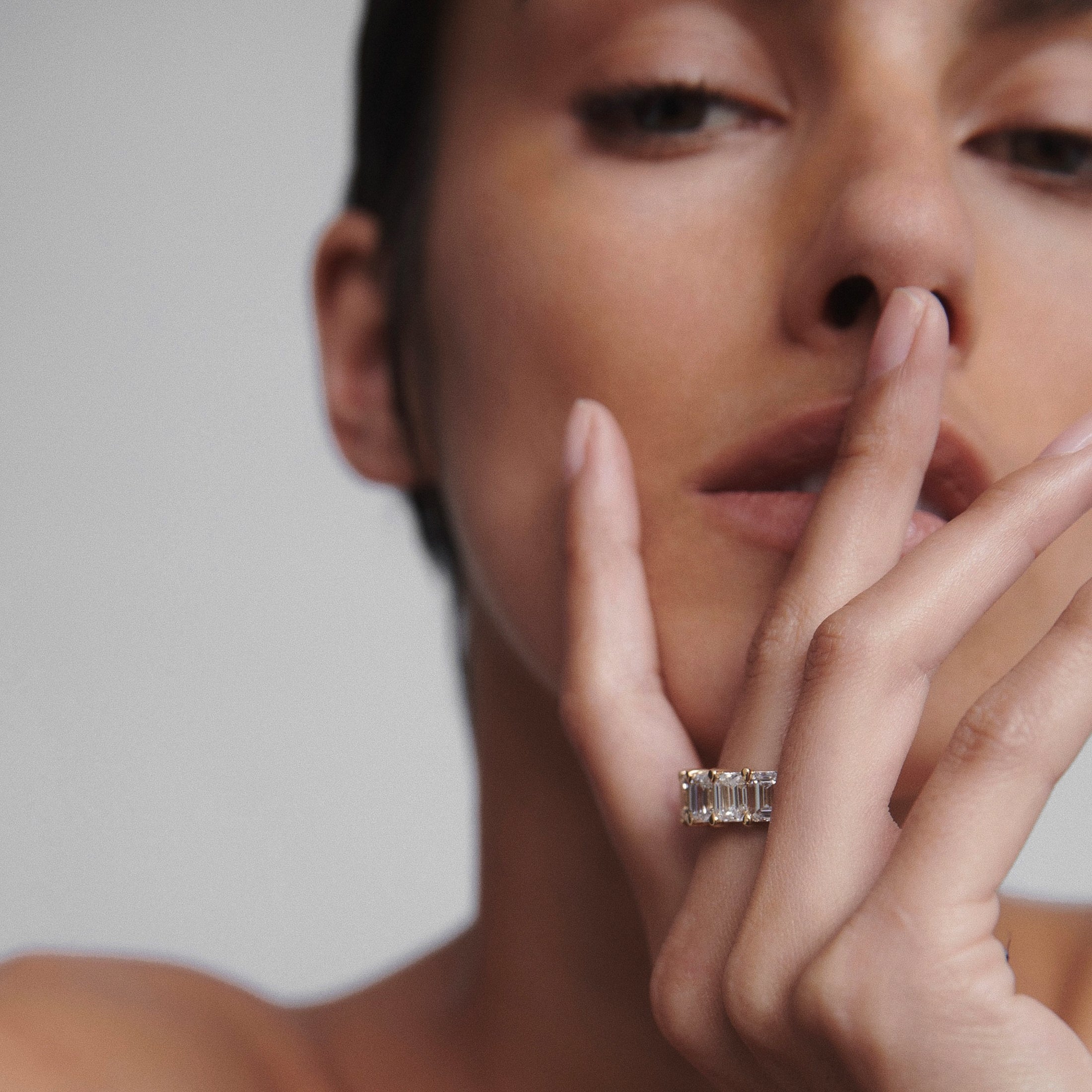 Close-up of a woman wearing a luxury emerald-cut diamond ring on her finger, softly touching her lips. High-end jewellery, elegance, sophistication.