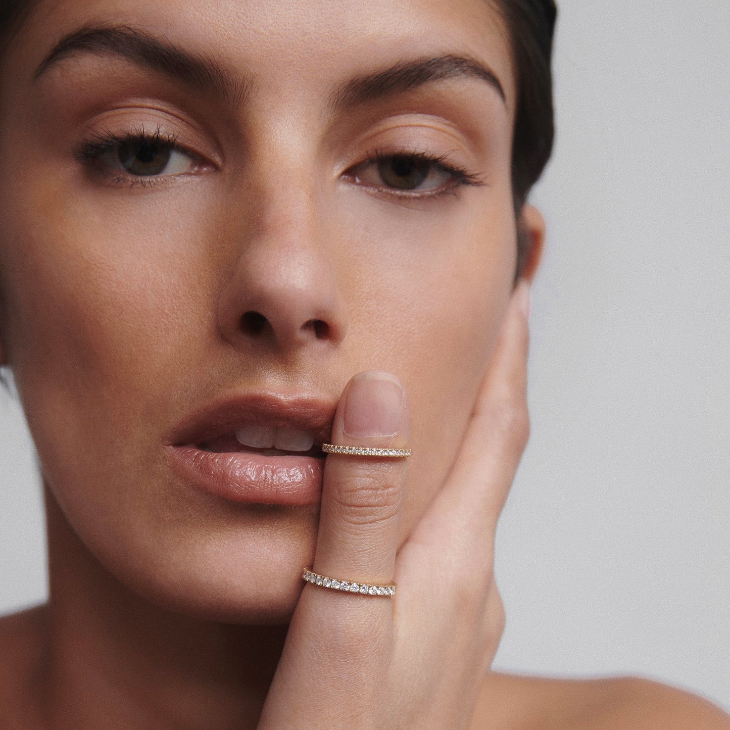 Woman with gold diamond rings on index and middle fingers, gently touching her face. Elegant, luxury jewellery close-up on minimalist backdrop.