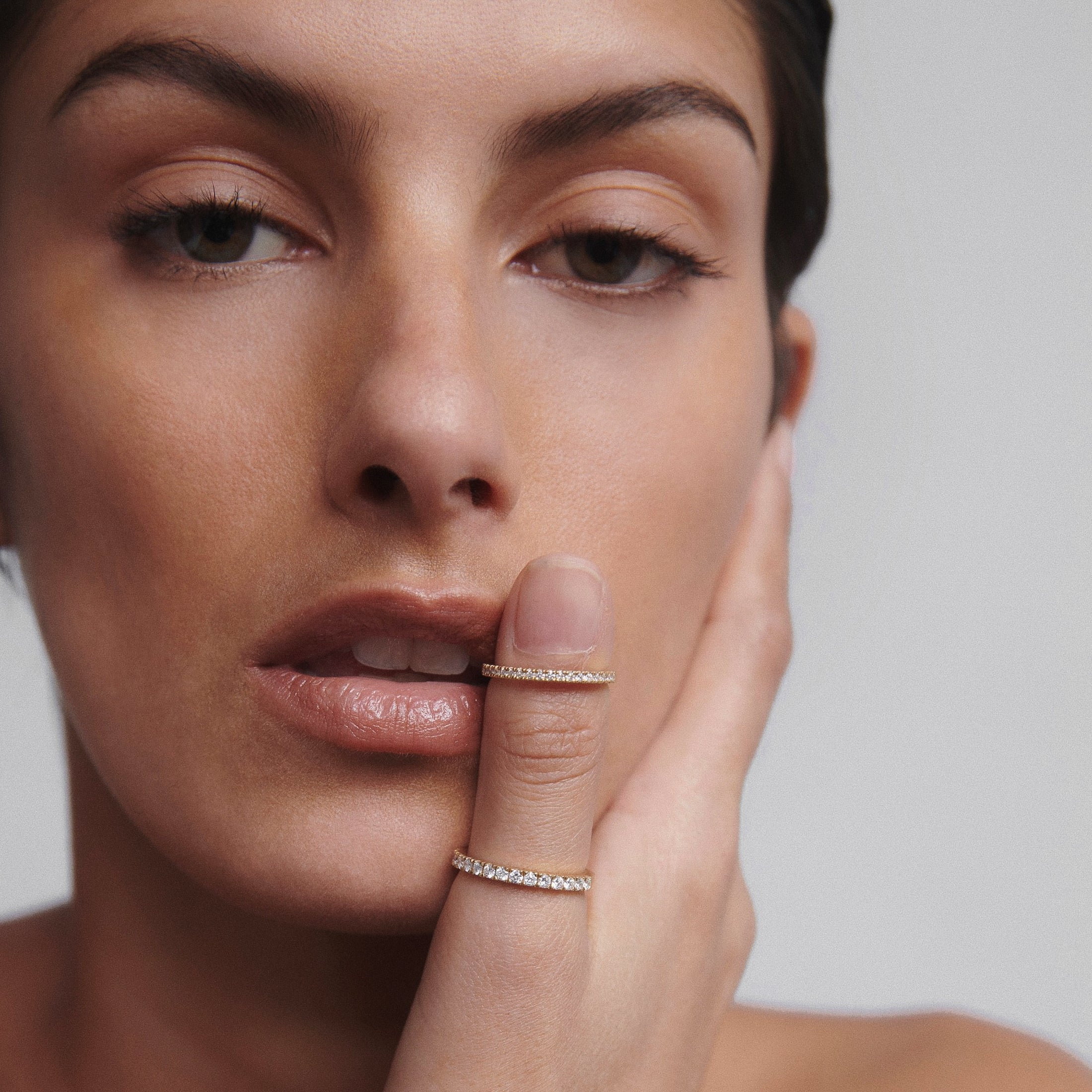 Woman with gold diamond rings on index and middle fingers, gently touching her face. Elegant, luxury jewellery close-up on minimalist backdrop.