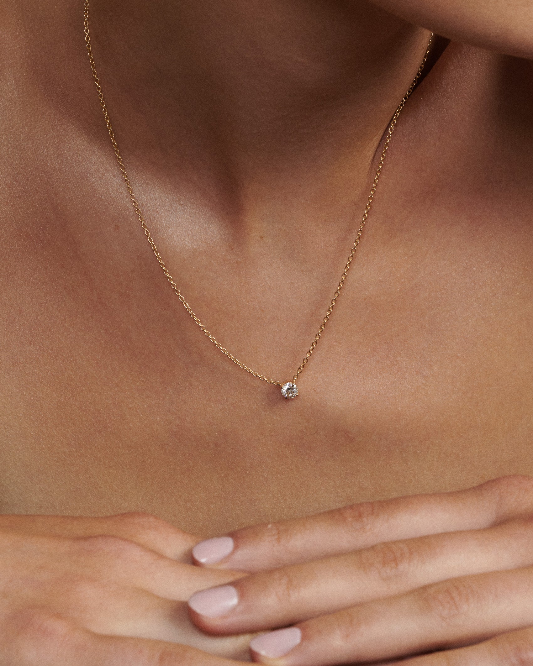 Gold necklace with a small diamond pendant on a person's neck [text overlay: Andrea wears the 0.5ct Round Diamond Necklace]