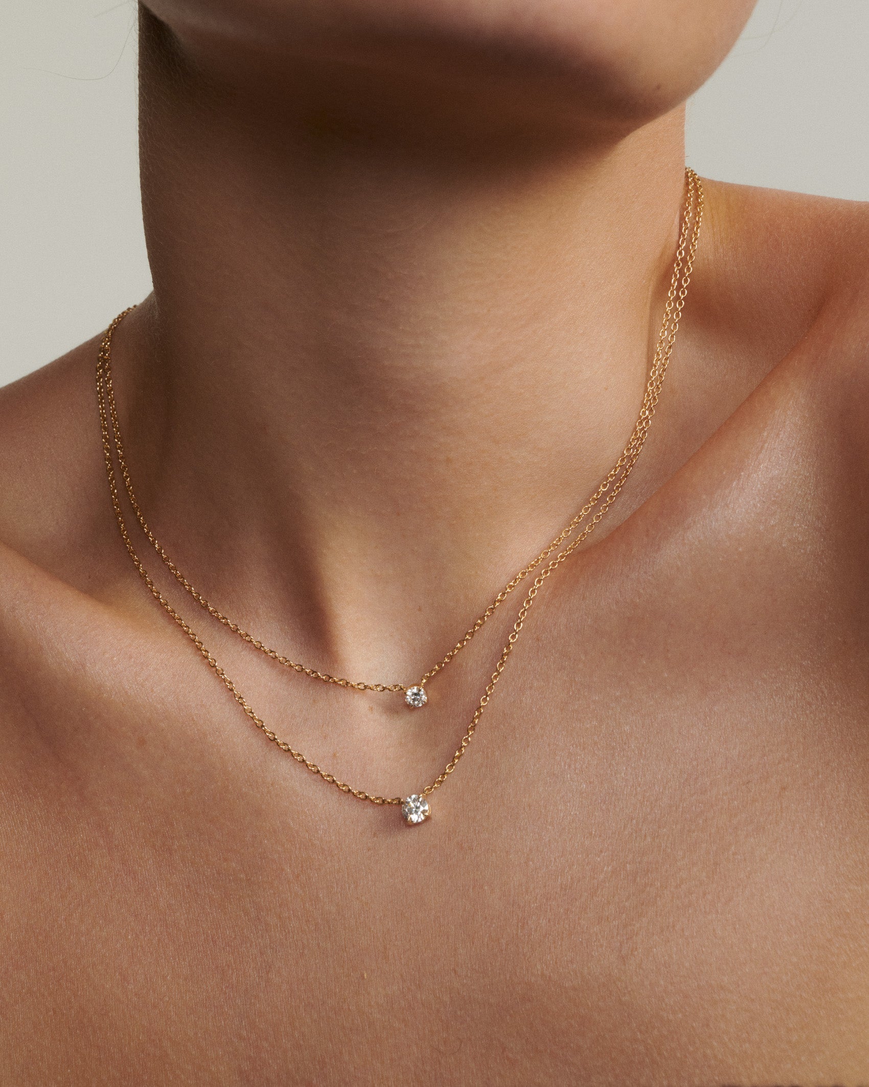 Gold necklace with small pendants on a person's neck against a neutral background [text overlay: Andrea wears the 0.25ct & 0.5ct Round Diamond Necklace]