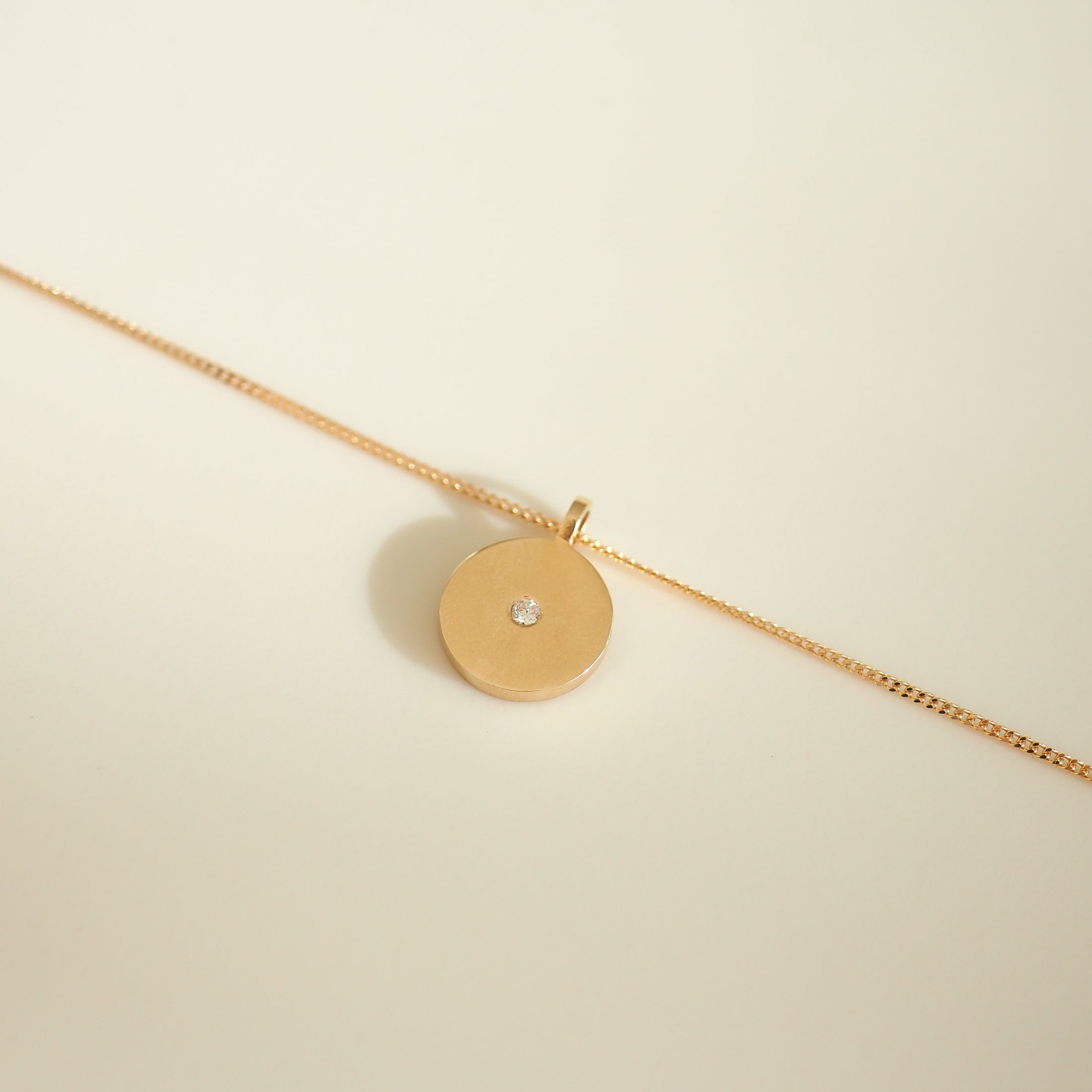 Gold necklace with a round pendant featuring a single diamond, displayed on a light background. Simple luxury jewellery design.