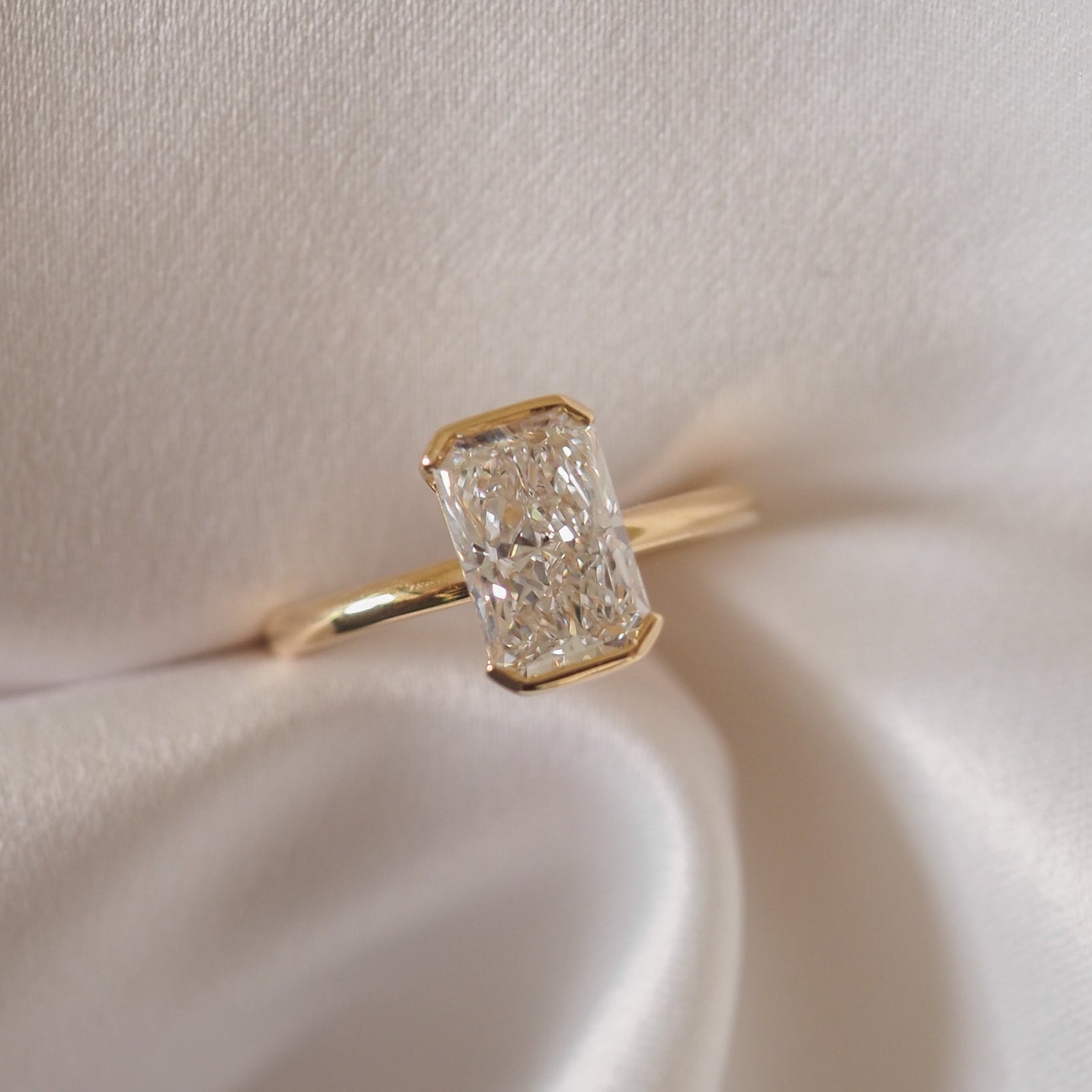 A radiant-cut diamond ring with a gold band, elegantly displayed on silky white fabric, showcasing luxury jewellery craftsmanship.