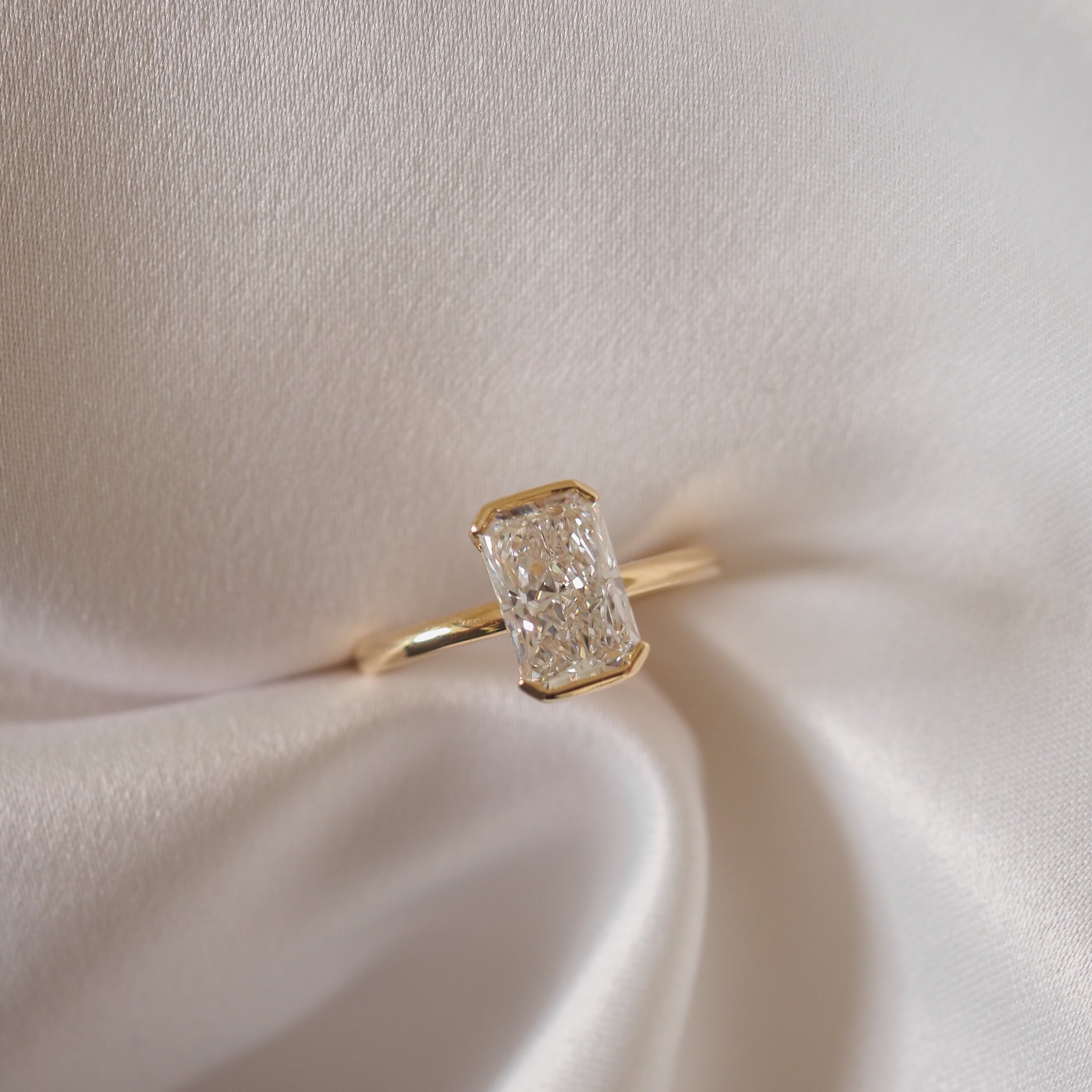 A radiant-cut diamond ring with a gold band, elegantly displayed on silky white fabric, showcasing luxury jewellery craftsmanship.
