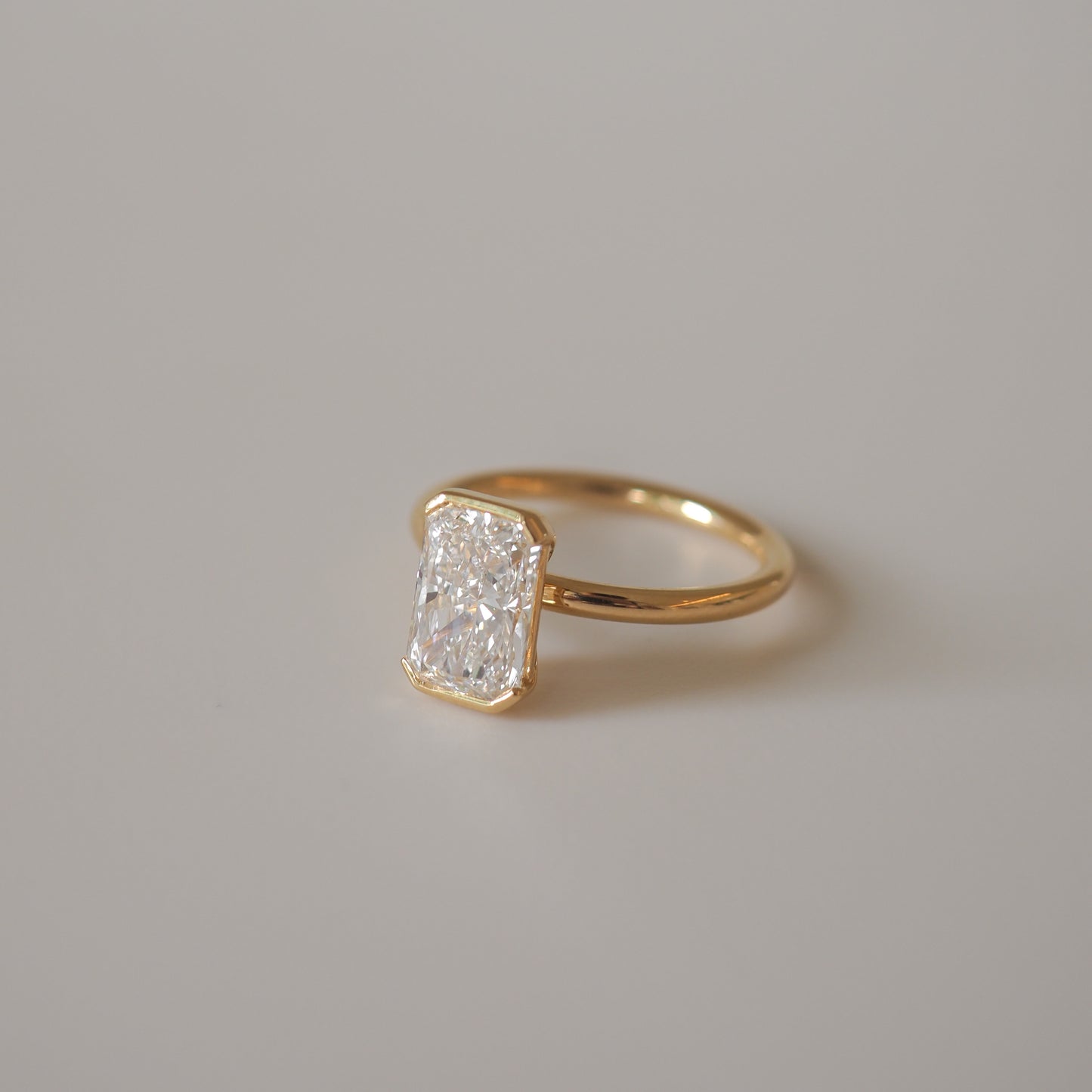 Gold ring with a radiant-cut diamond in a minimalist setting, displayed on a neutral background. Luxurious and elegant jewellery piece.