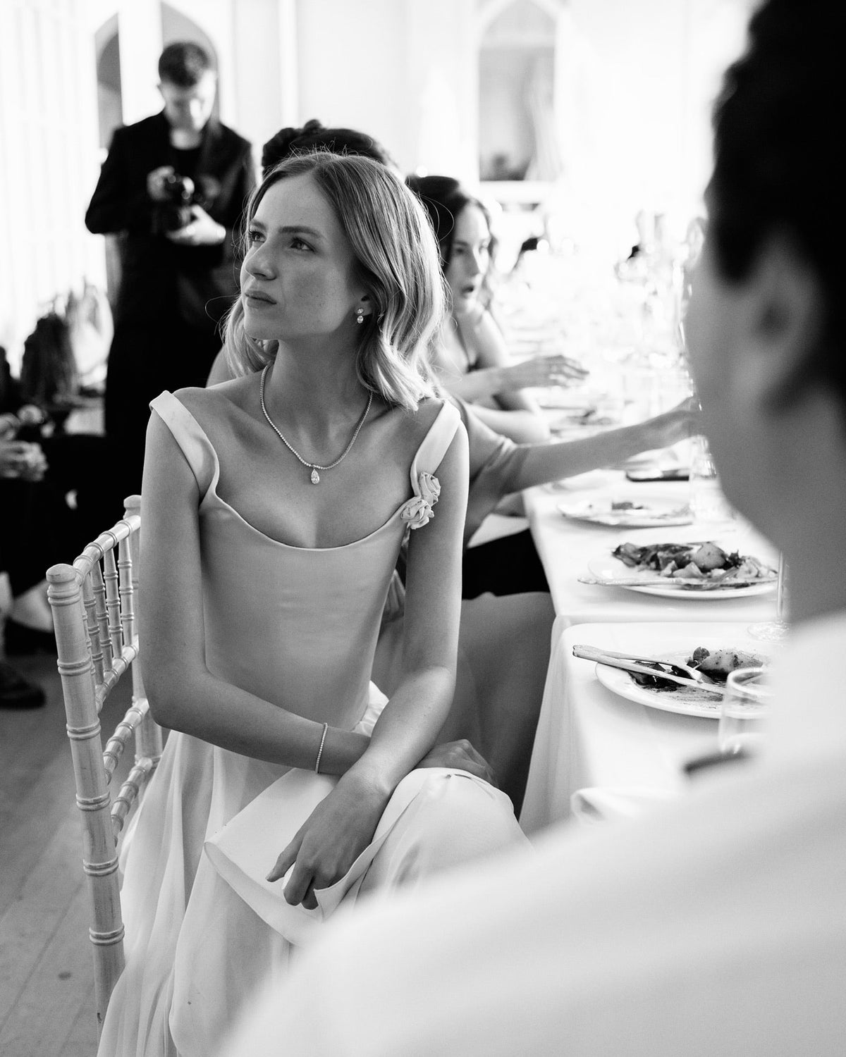 A black and white photograph of a woman in a sleeveless, scoop-neck white dress sitting at a reception table, looking off-camera. The image has a shallow depth of field with a blurred foreground figure and other guests in a bright indoor venue.