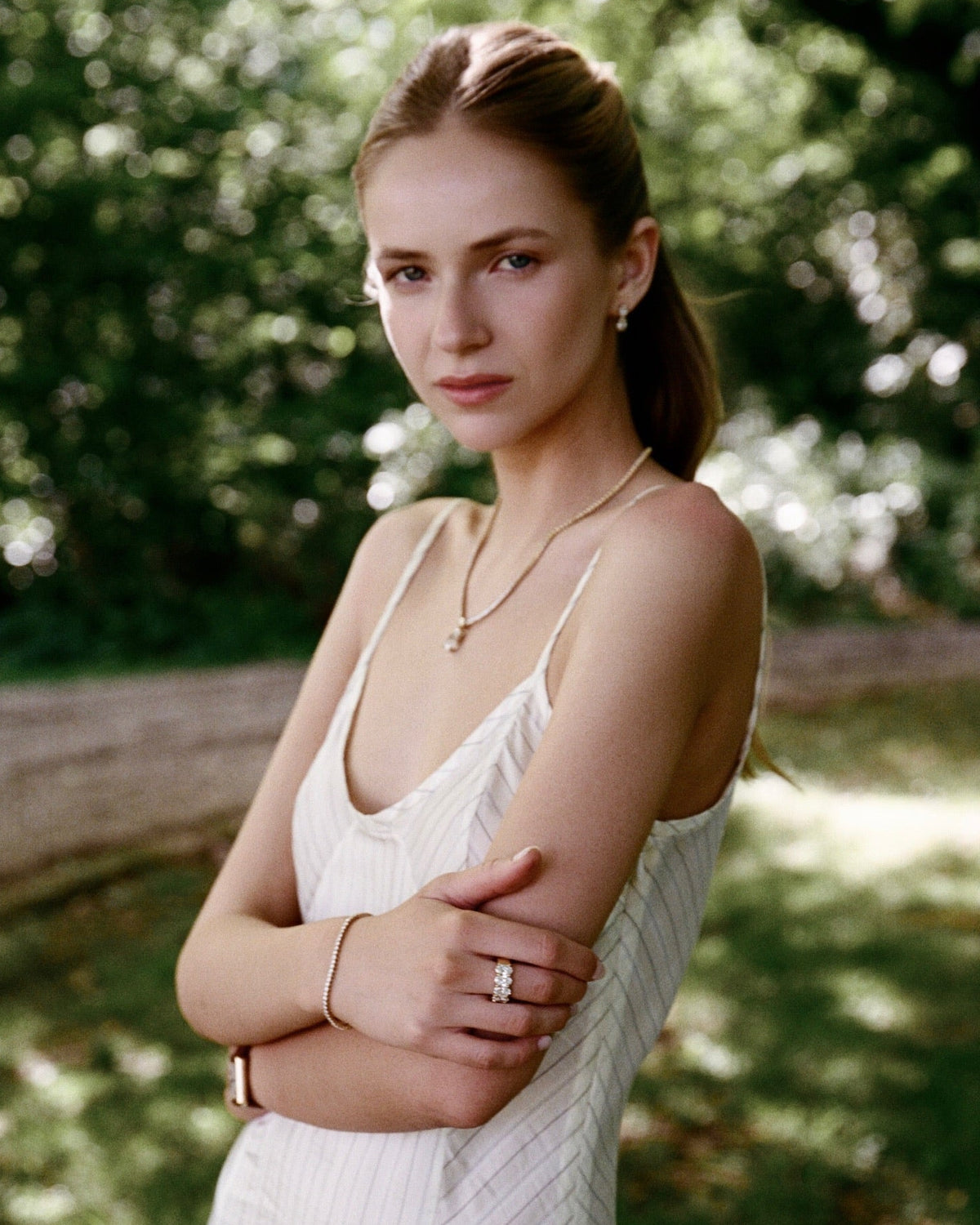 A woman stands outdoors in a sunlit wooded setting, wearing a white sleeveless textured dress and gold jewelry, including a necklace and ring.