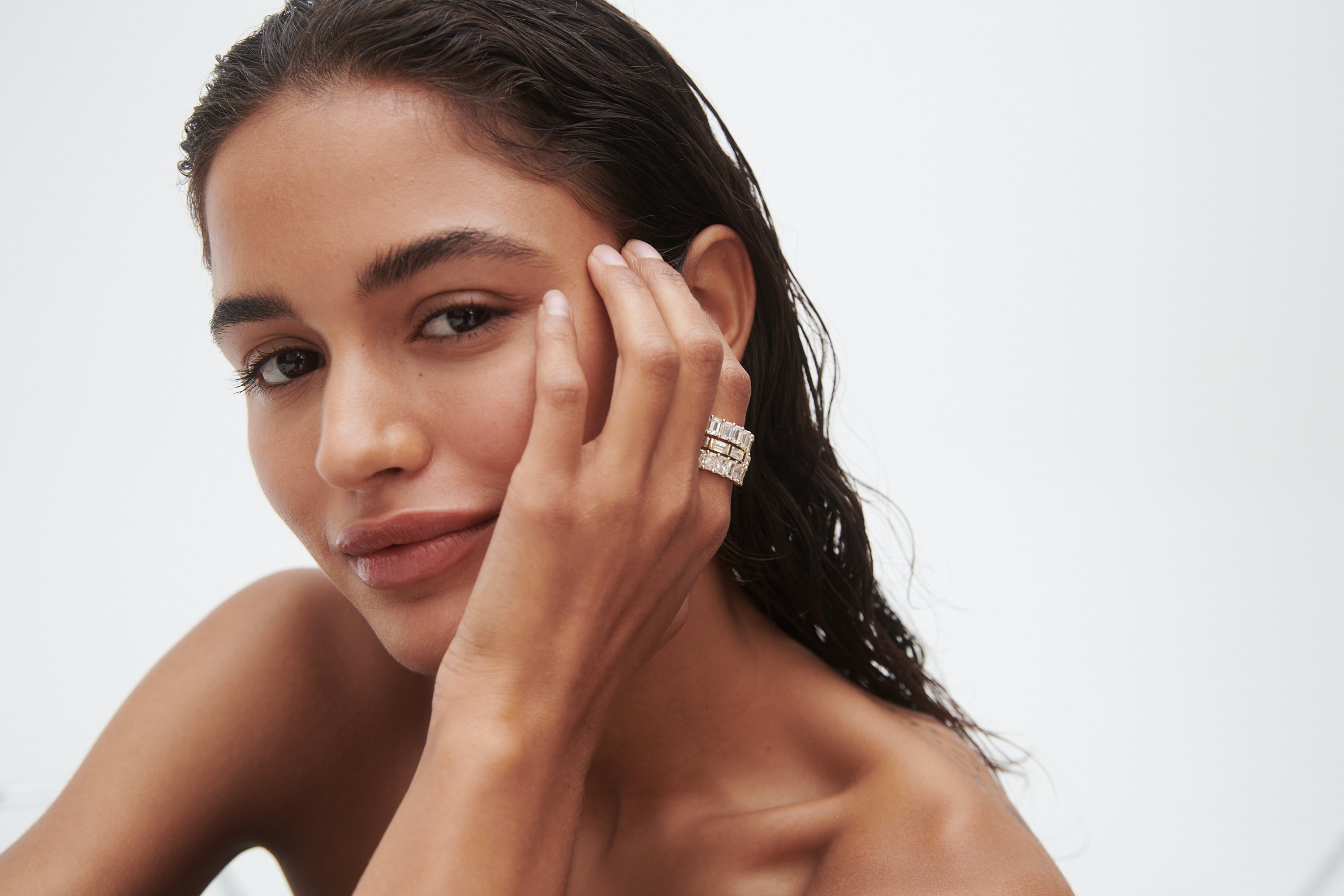 Woman with wet hair poses, showcasing elegant stacked rings on her hand against her face. Luxurious jewellery photo with minimalist style.
