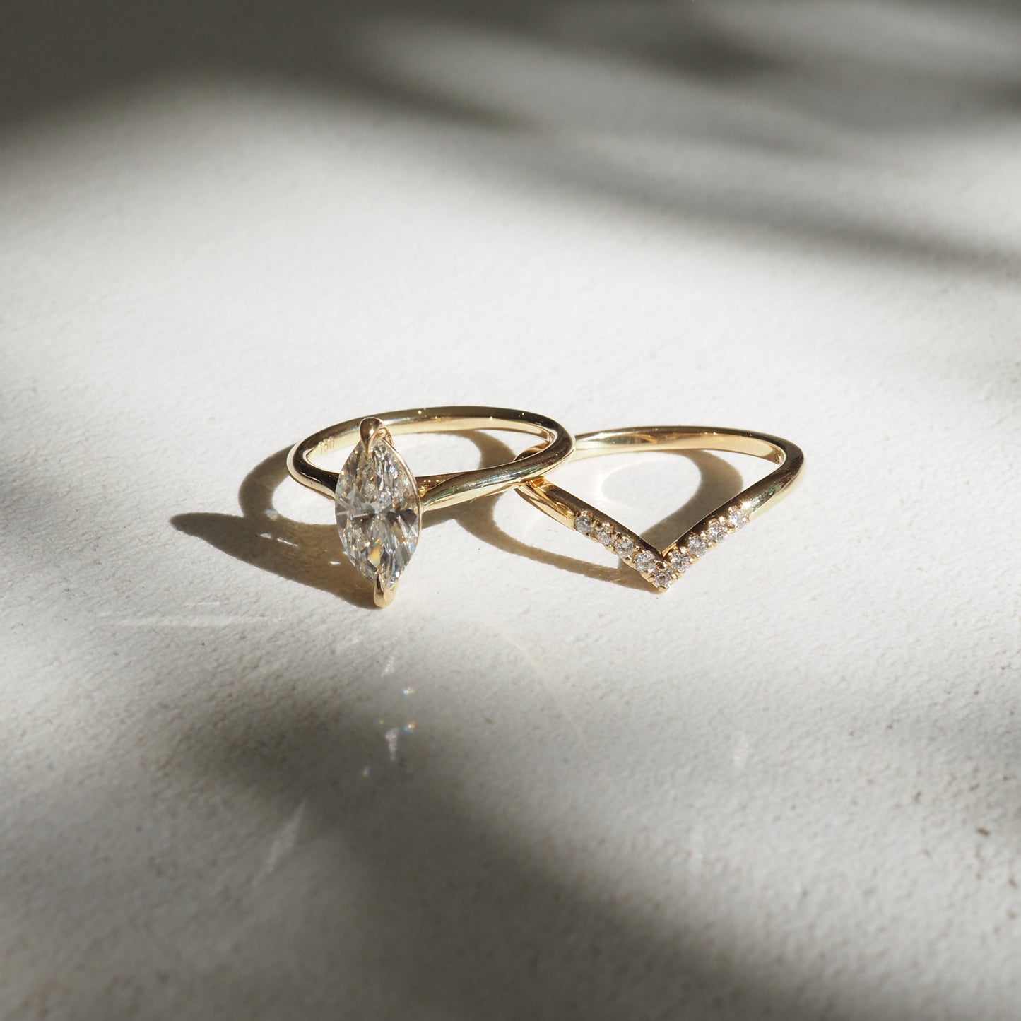 Two gold rings on a sunlit surface: one with a marquise diamond and another with a V-shaped design adorned with small diamonds. Luxury jewellery.
