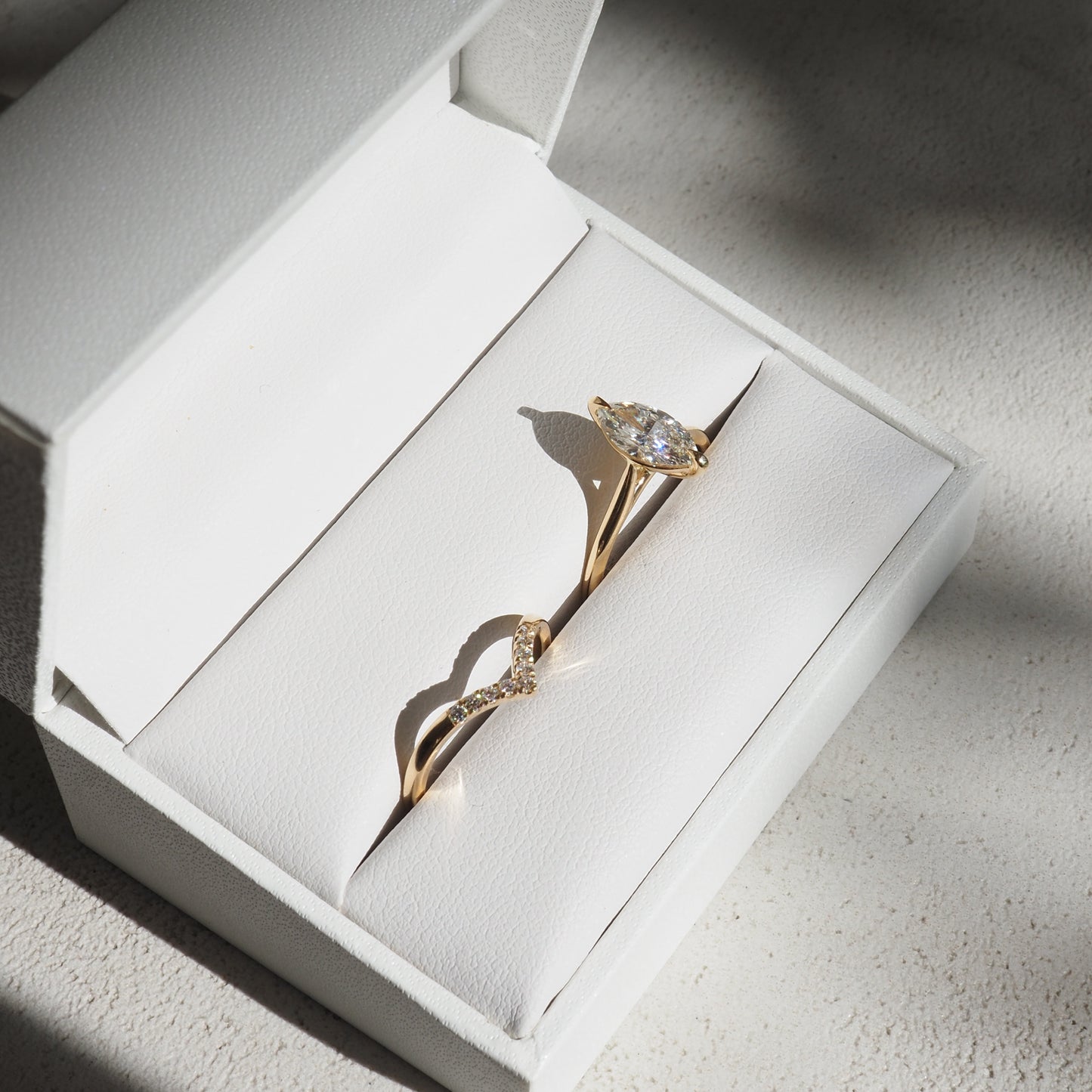 Gold engagement ring with marquise diamond in a white jewellery box. Sunlight casts soft shadows on elegant design.