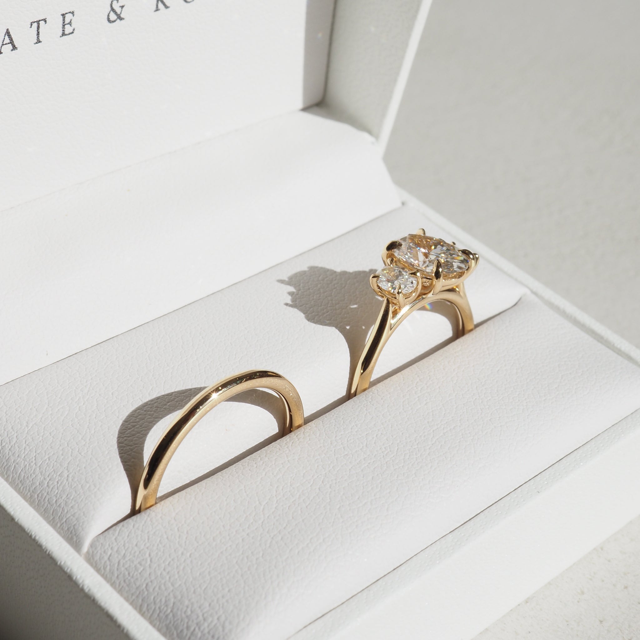 Gold diamond engagement and wedding rings set in a white jewellery box, illuminated by soft natural light, showcasing luxury craftsmanship.