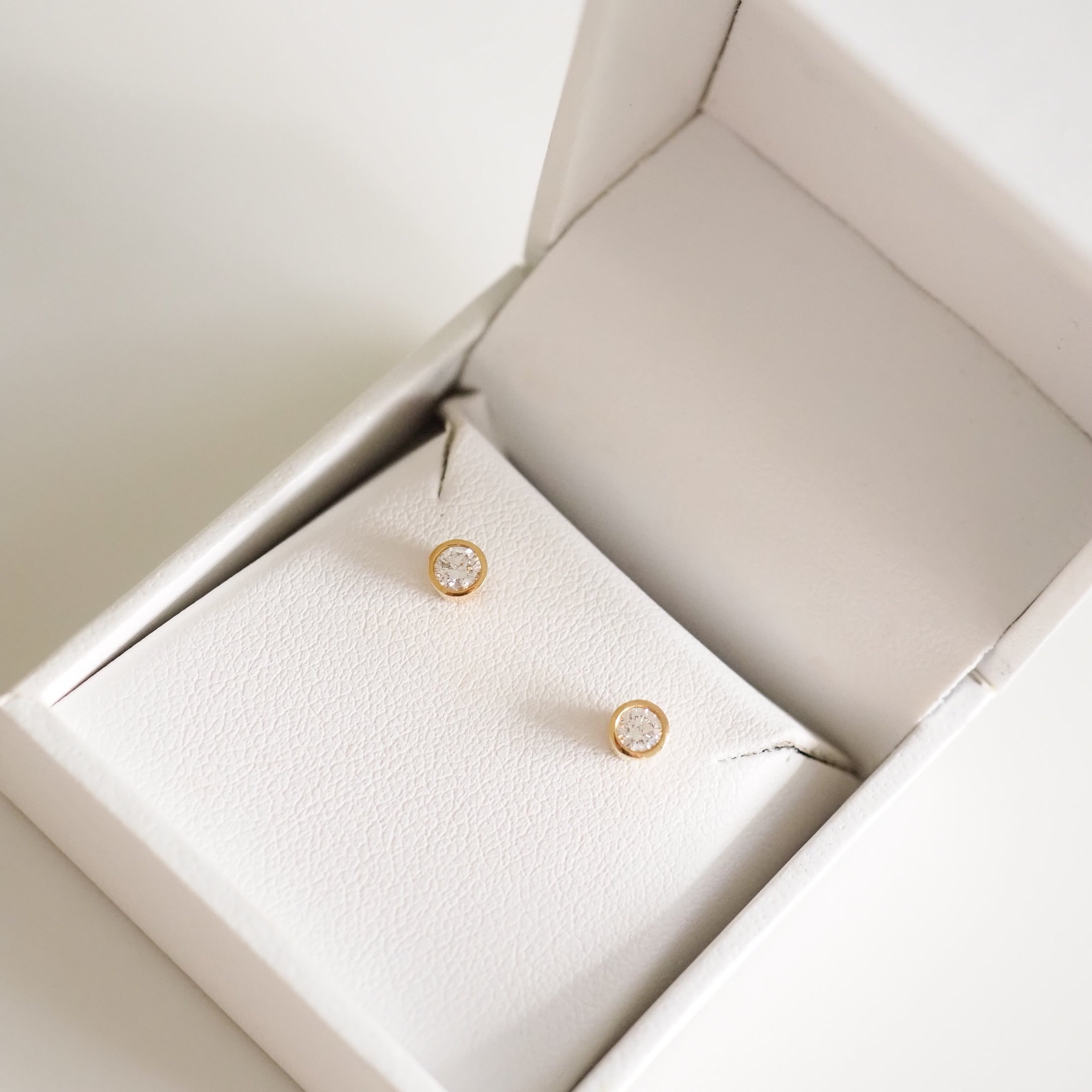 Gold bezel-set diamond stud earrings in a white jewellery box, showcasing elegant luxury and craftsmanship.