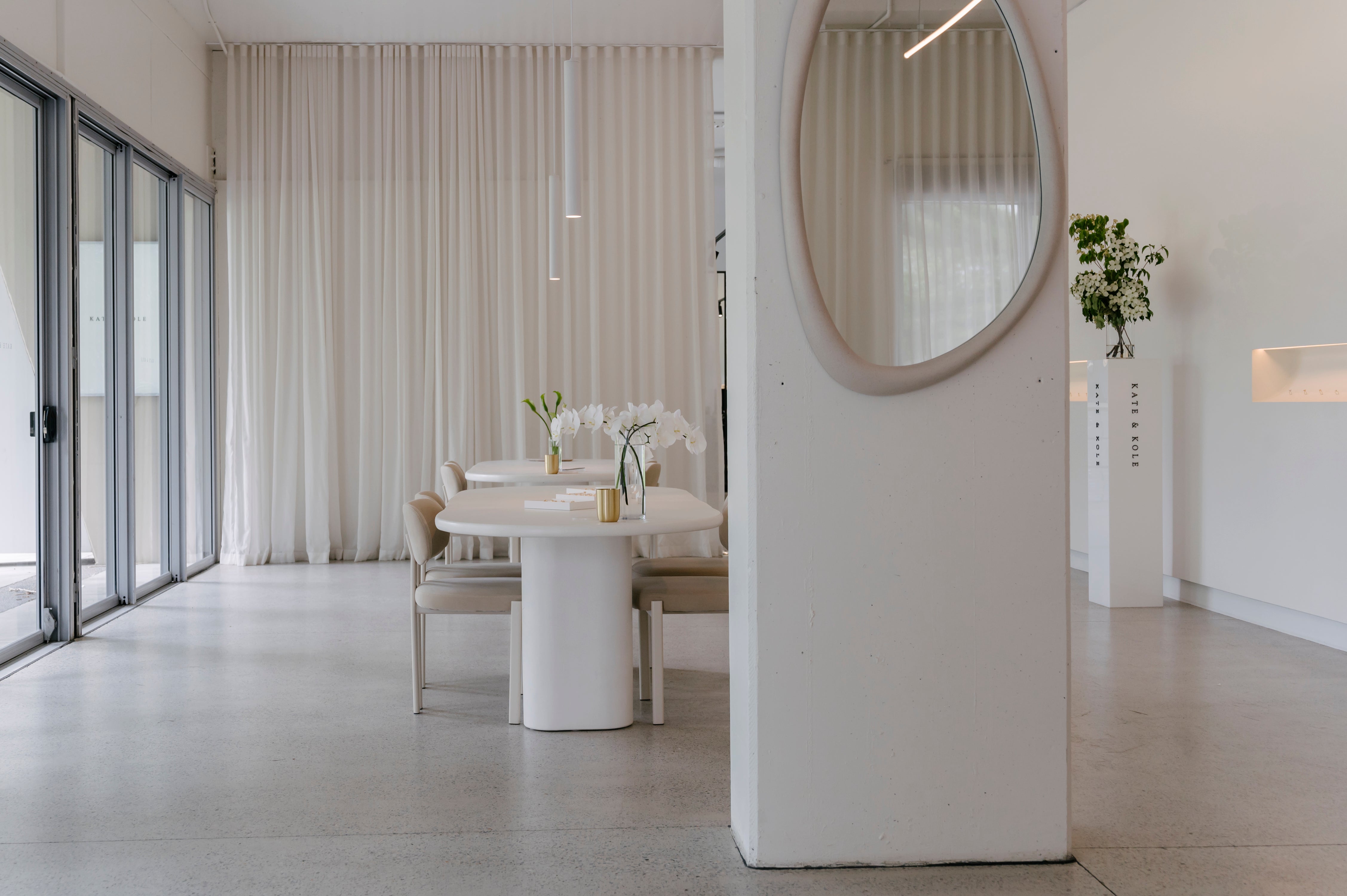 Minimalist luxury jewellery showroom with white walls, round table, elegant chairs, soft curtains, and a decorative mirror. Modern and serene space.