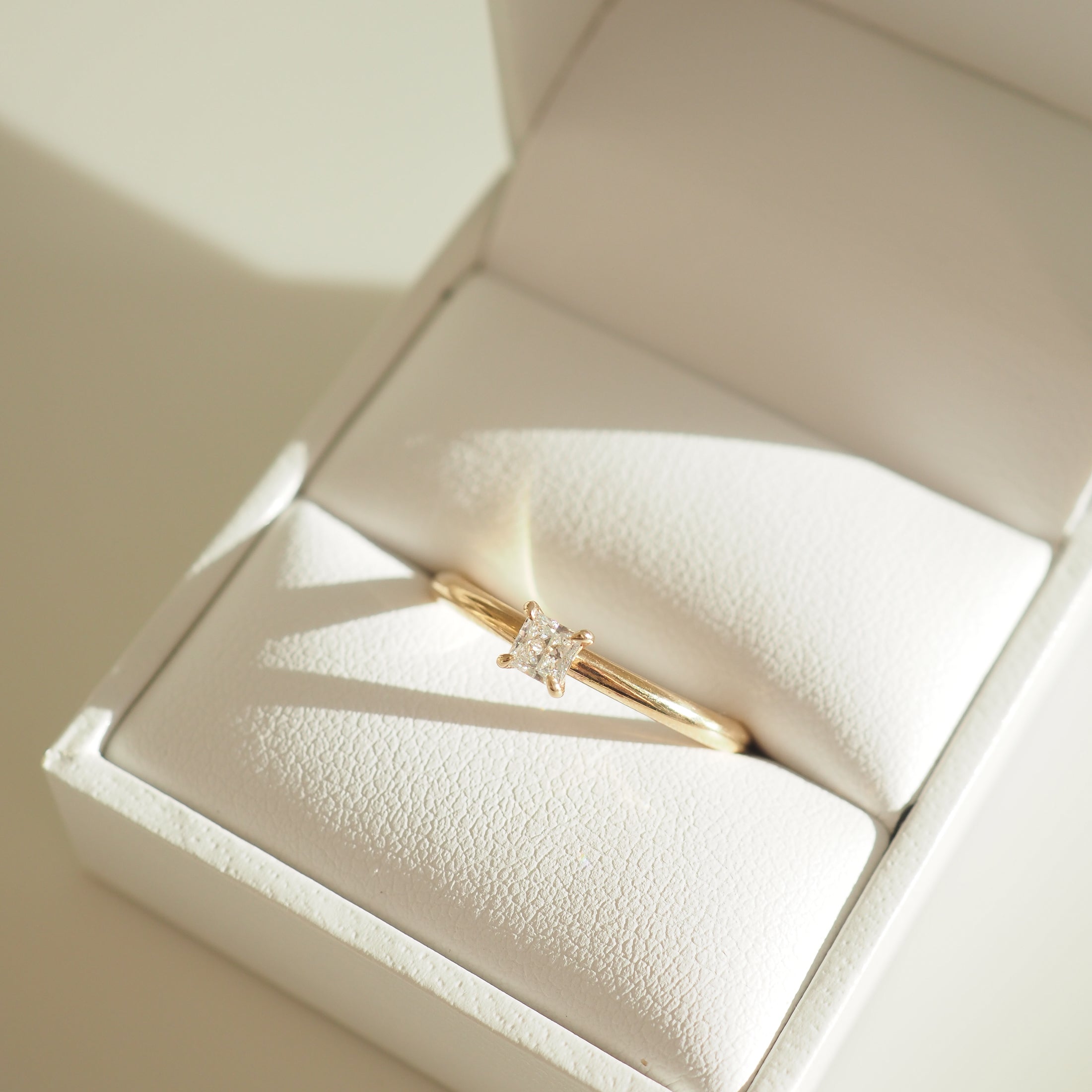 Gold solitaire engagement ring with square-cut diamond in a white jewellery box. Sunlight casts soft shadows, highlighting luxury design.