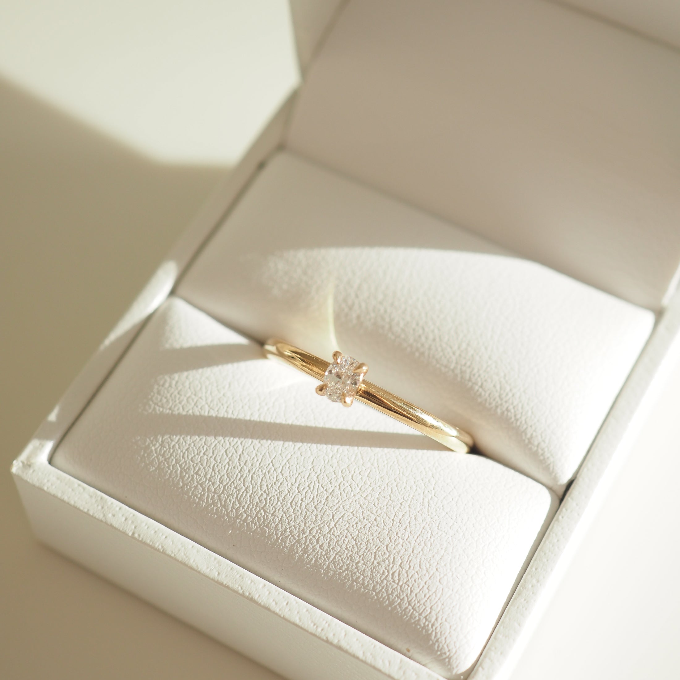 Gold ring with a solitaire diamond, elegantly displayed in a white jewellery box. Sunlight casts soft shadows on the fabric. Luxurious and refined.