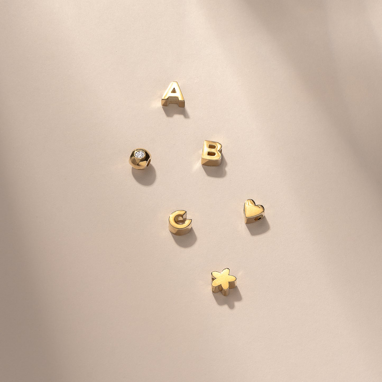 Gold alphabet charms and symbols, including letters A, B, C, a heart, a star, and a diamond stud, elegantly arranged on a light surface.