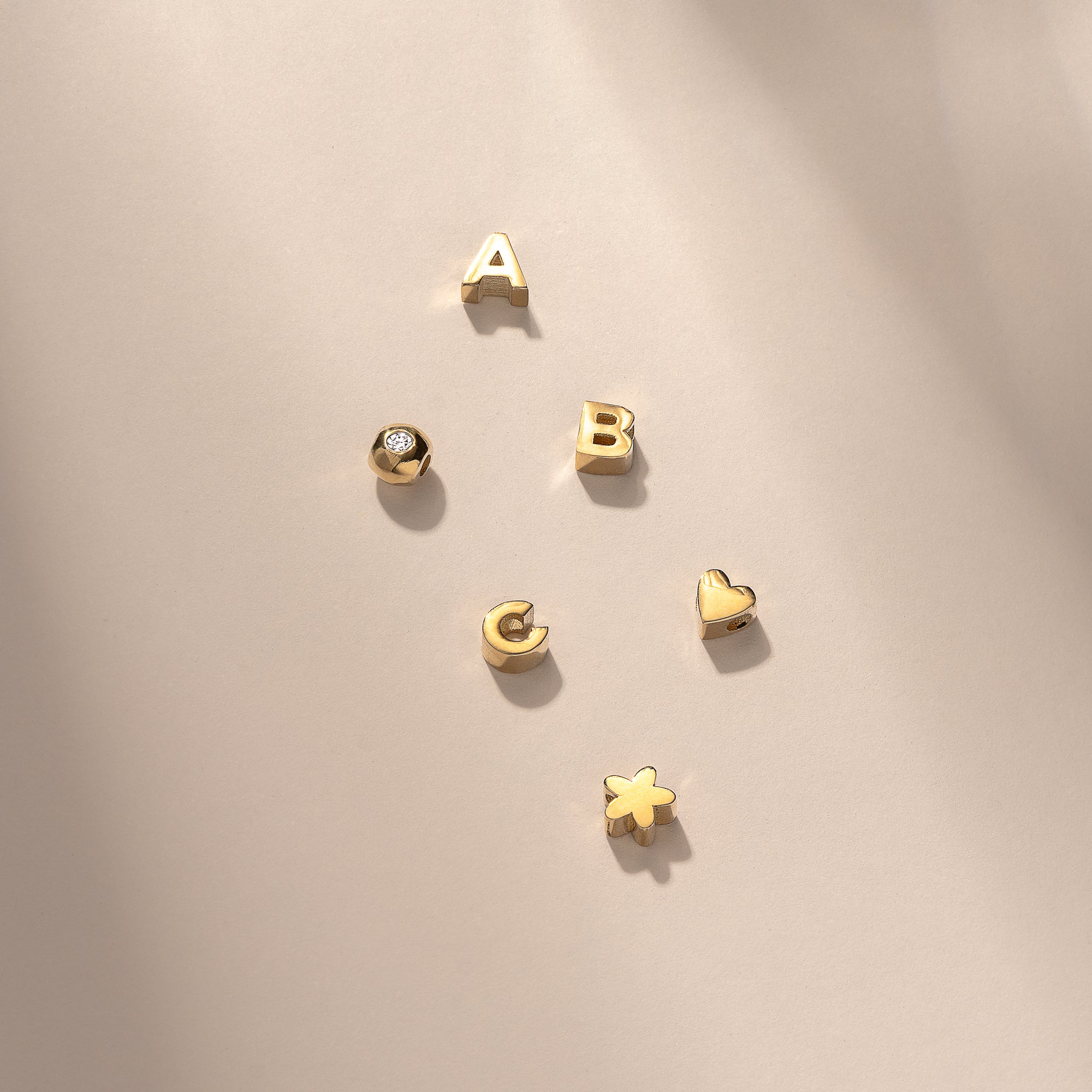 Gold alphabet charms and symbols, including letters A, B, C, a heart, a star, and a diamond stud, elegantly arranged on a light surface.