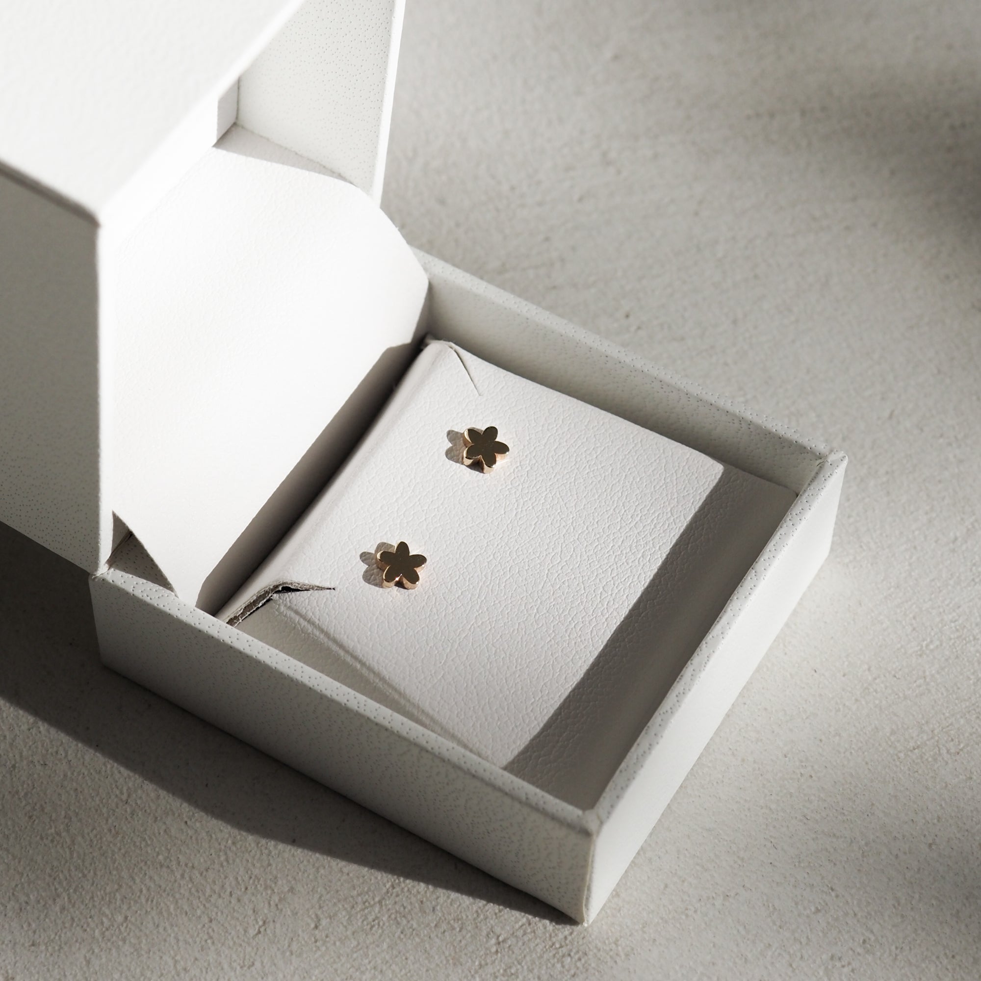 Gold flower-shaped stud earrings in a white jewellery box on a light grey surface, softly lit with shadows.