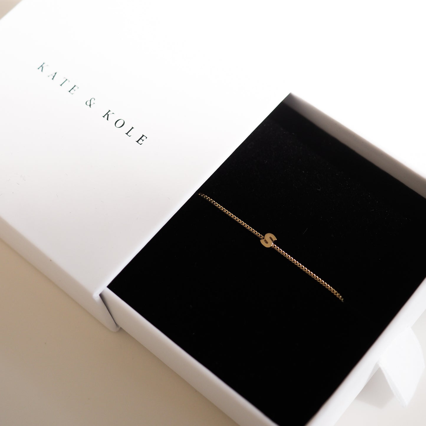 Gold bracelet with 'S' charm in a black velvet jewellery box by Kate & Kole, set against a white background.