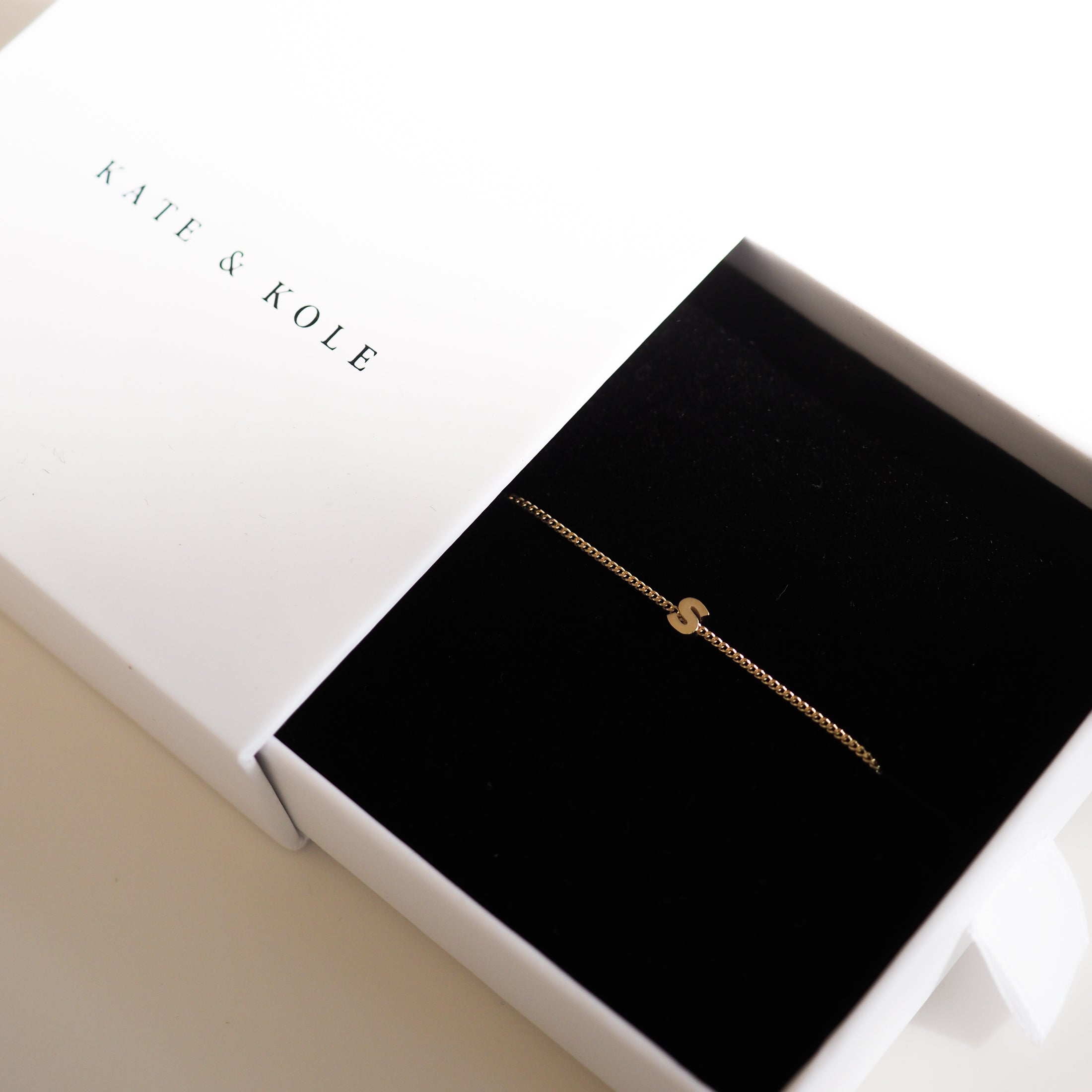 Gold bracelet with 'S' charm in a black velvet jewellery box by Kate & Kole, set against a white background.