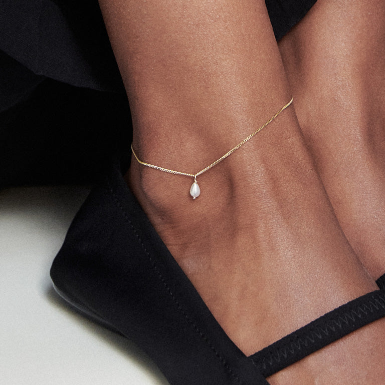 Elegant gold anklet with a teardrop pearl pendant on a person's ankle, wearing black shoes. Perfect for high-end jewellery enthusiasts.