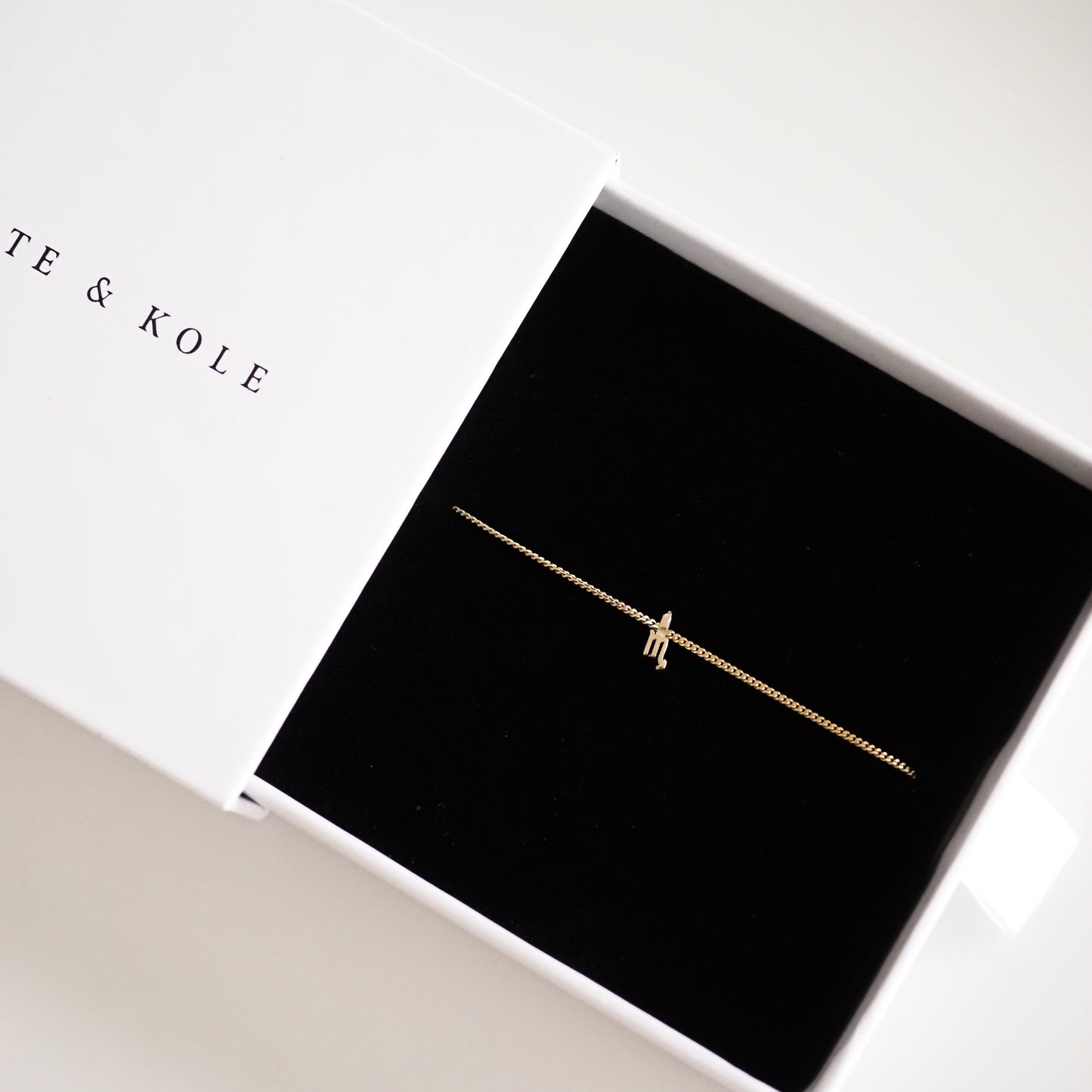 Delicate gold bracelet with a tiny charm in a sleek black jewellery box, partially covered by a white lid.