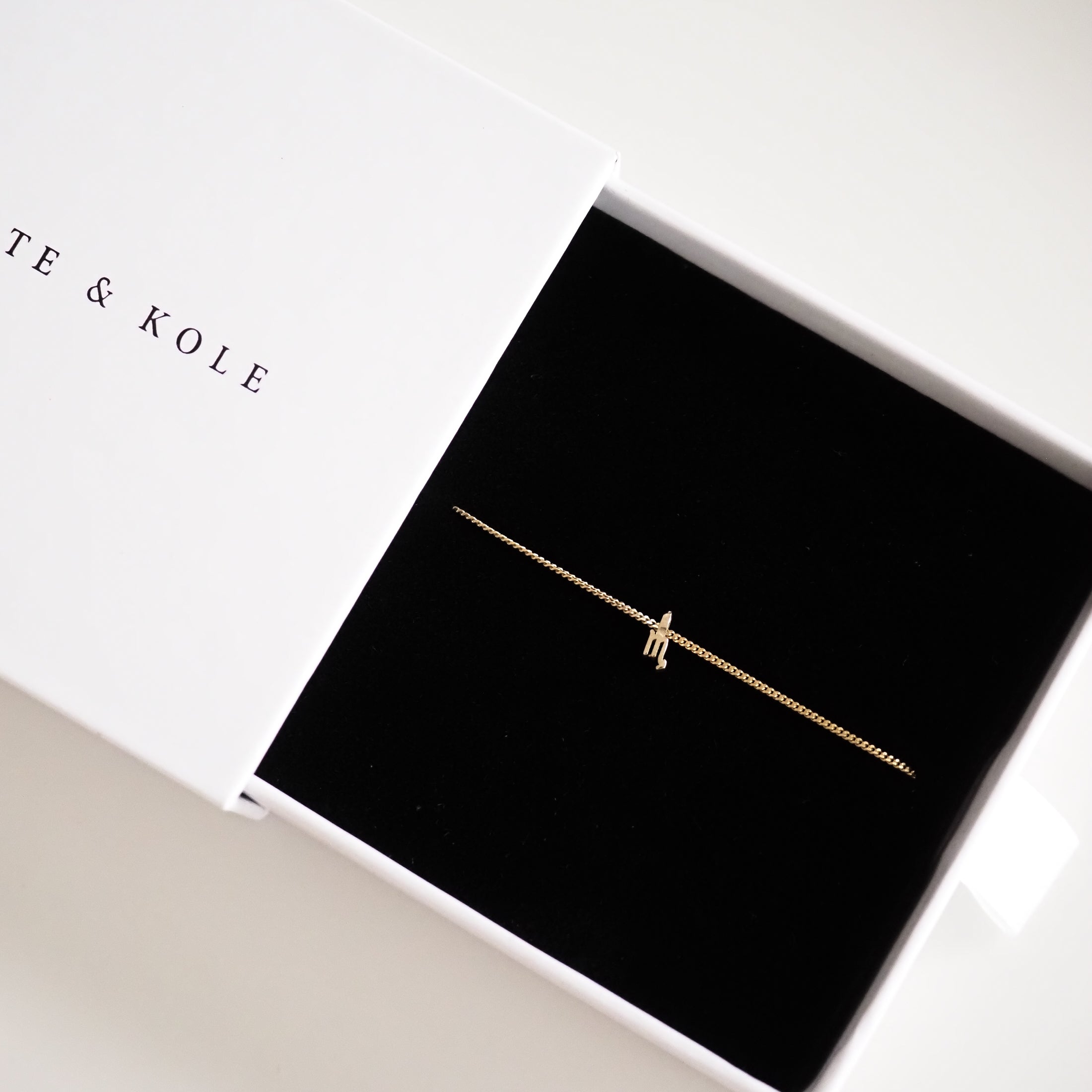 Delicate gold bracelet with a tiny charm in a sleek black jewellery box, partially covered by a white lid.