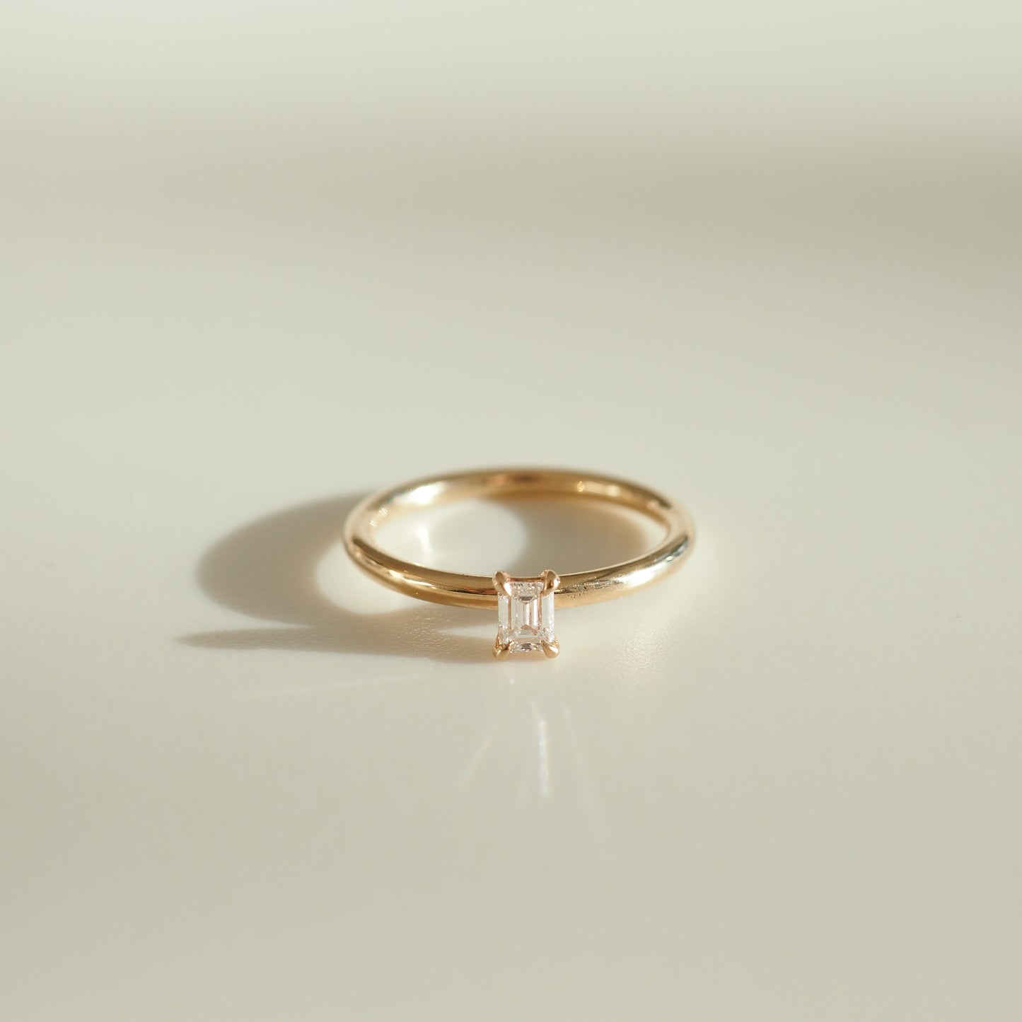 Elegant gold ring featuring a rectangular-cut diamond, displayed on a light-toned surface, casting a soft shadow.