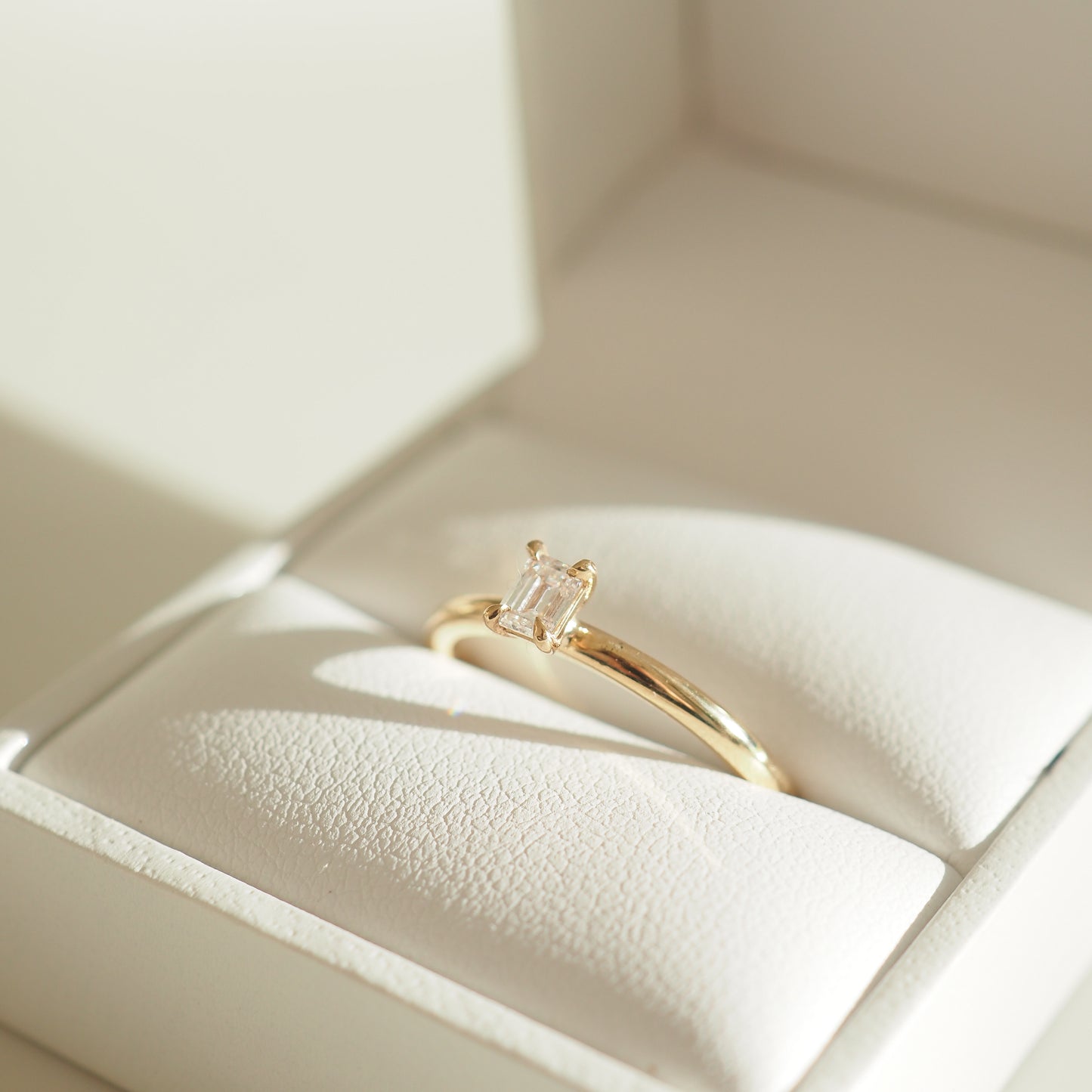 Gold ring with a rectangular diamond in an elegant white jewellery box, captured under soft sunlight.