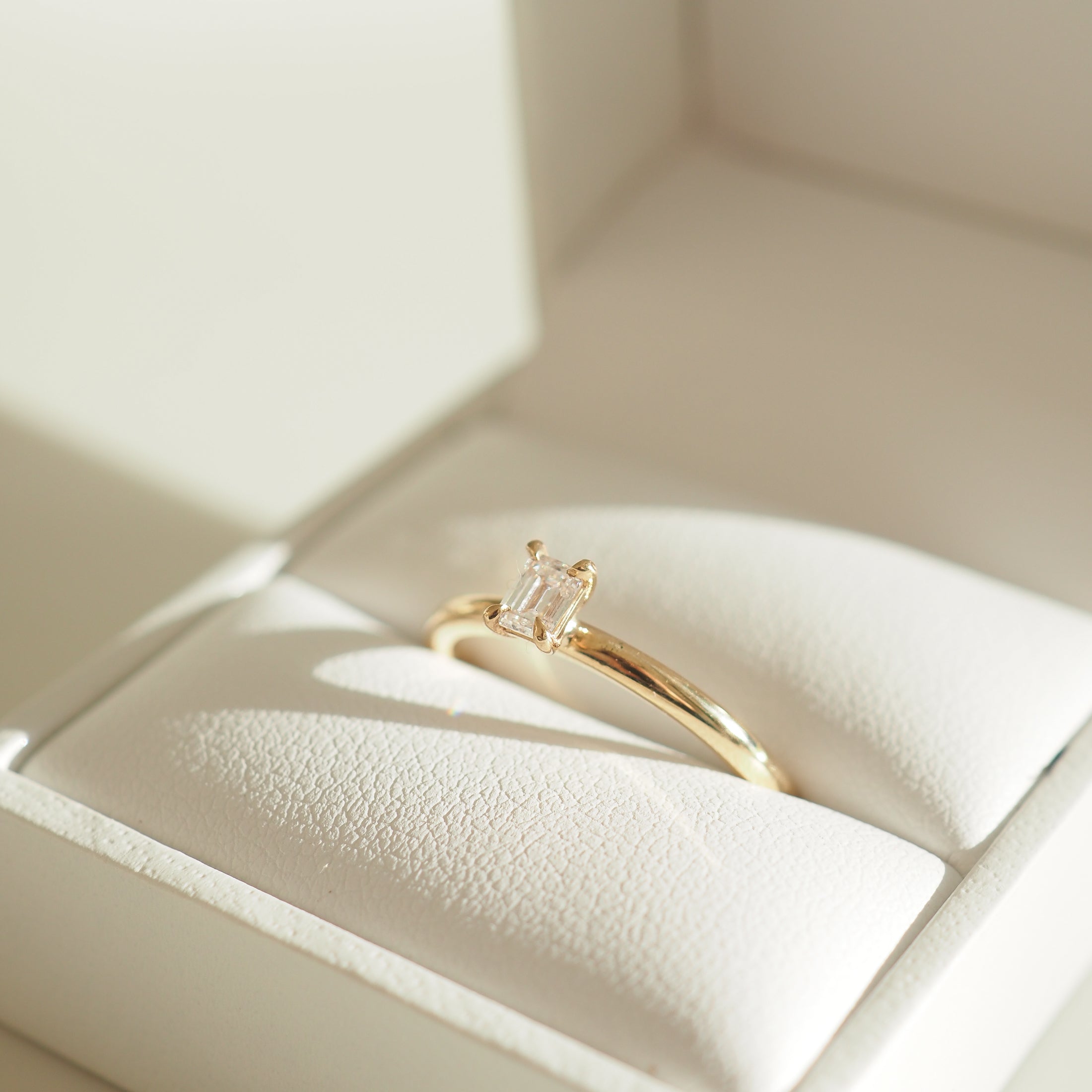 Gold ring with a rectangular diamond in an elegant white jewellery box, captured under soft sunlight.