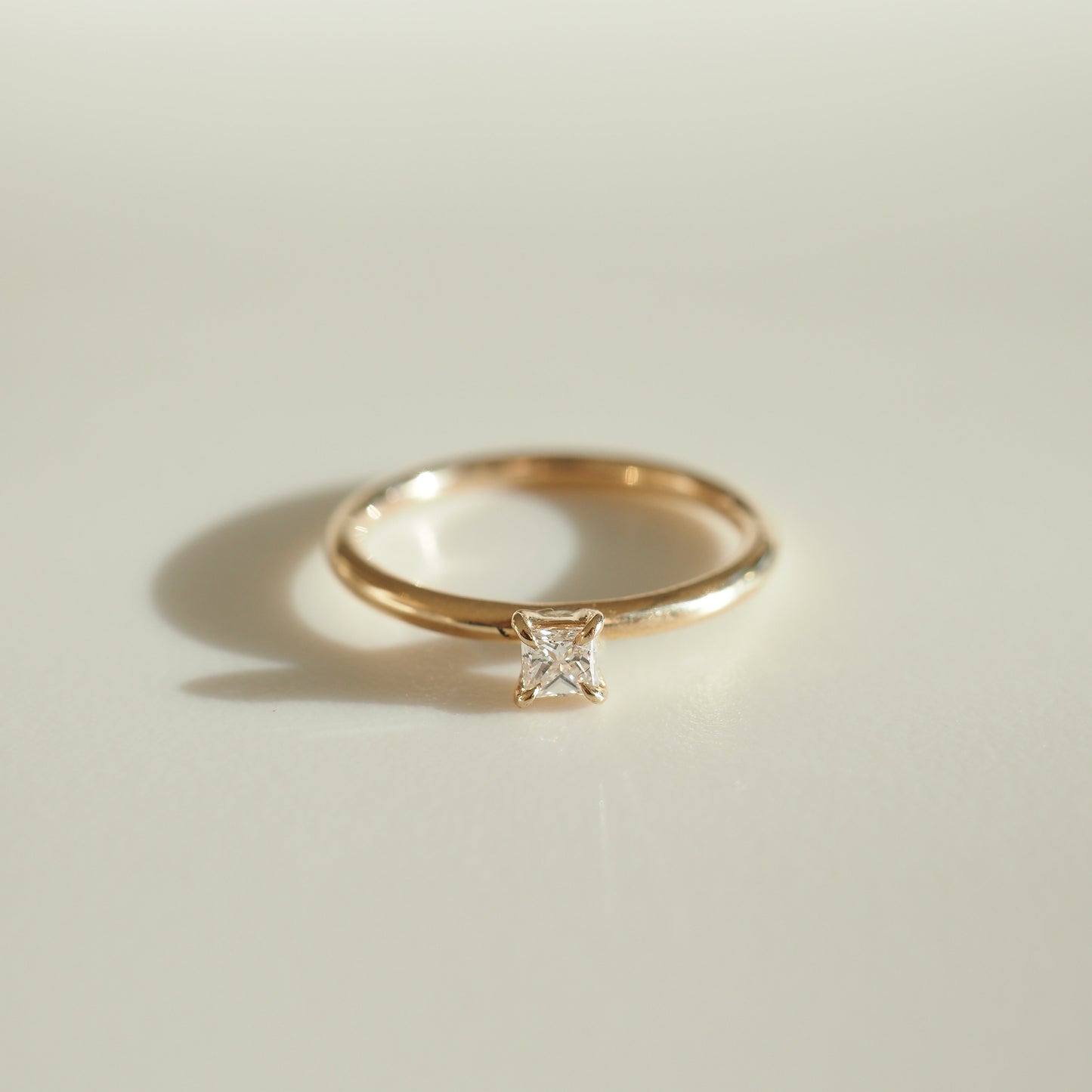 Gold ring with a square-cut diamond solitaire, elegantly displayed on a light surface, casting a soft shadow. Luxury jewellery.