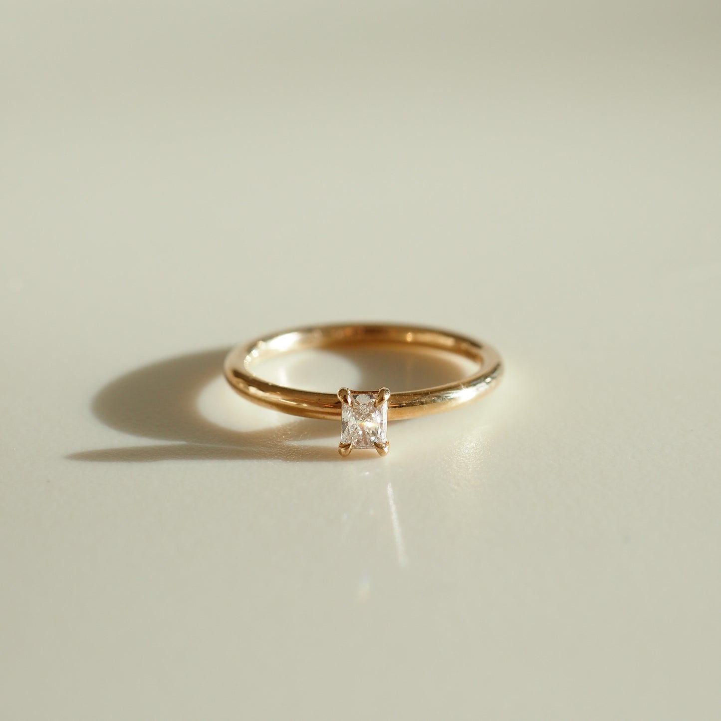 Gold ring with a princess-cut diamond set in a simple, elegant band, casting a shadow on a light surface. Perfect for luxury jewellery lovers.