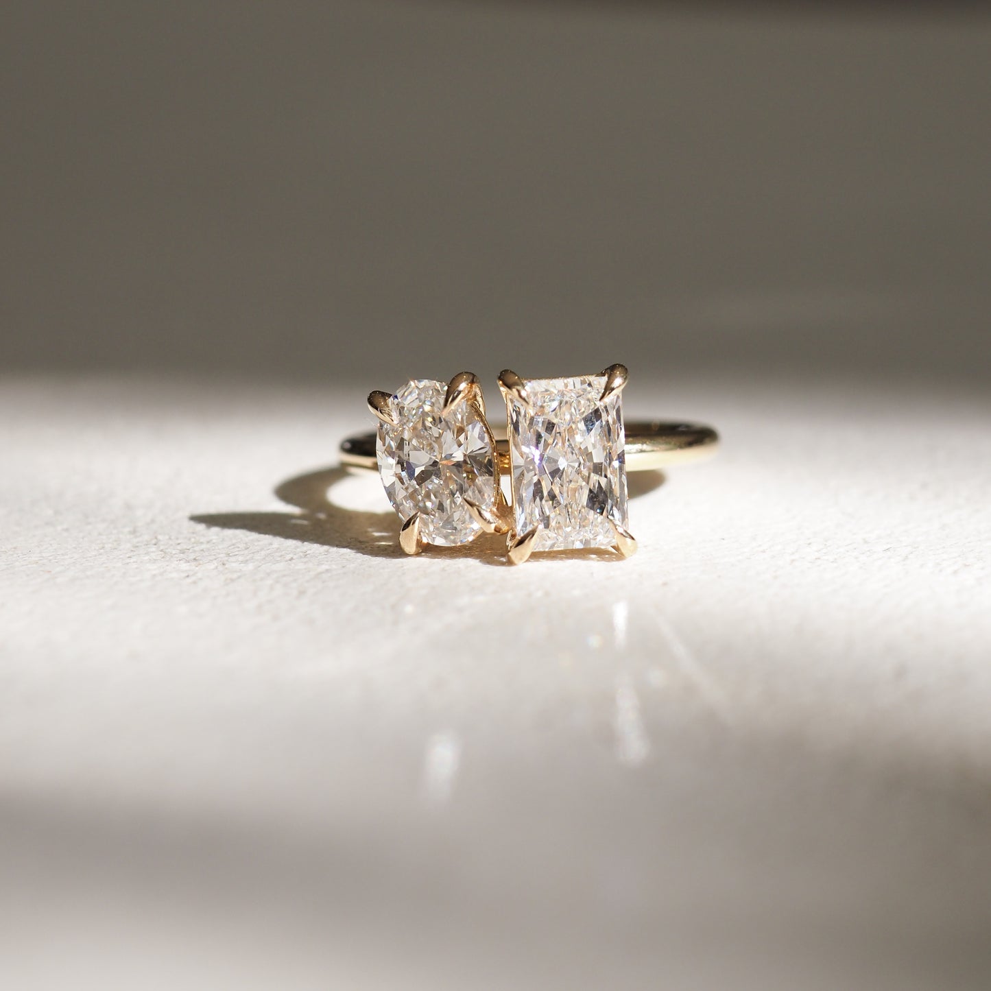 A gold ring with one radiant-cut and one oval diamond, catching sunlight on a soft surface, showcasing elegant luxury jewellery design.