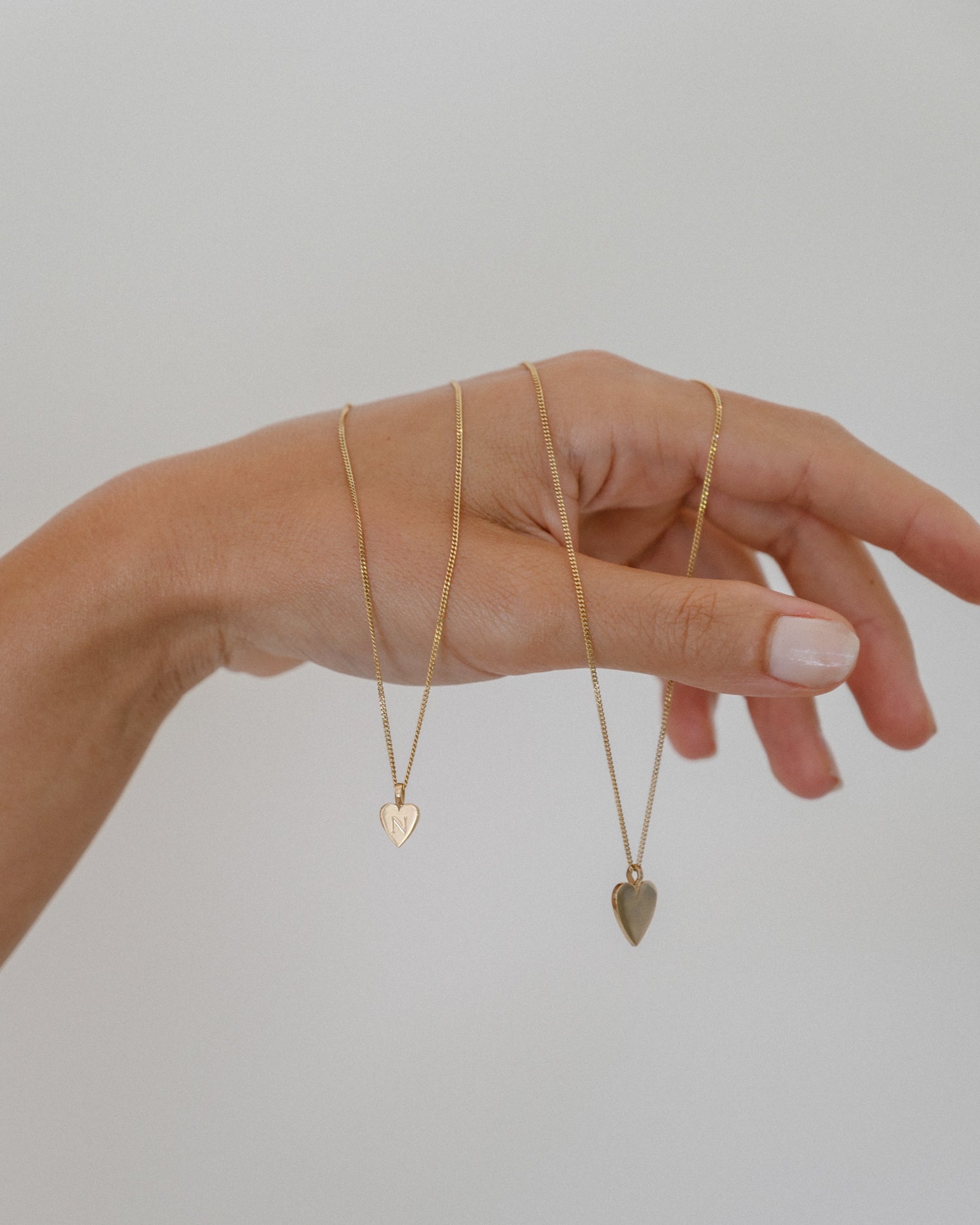 Hand elegantly holding two gold heart necklaces, showcasing luxury jewellery. Neutral background highlights the delicate, high-end design.