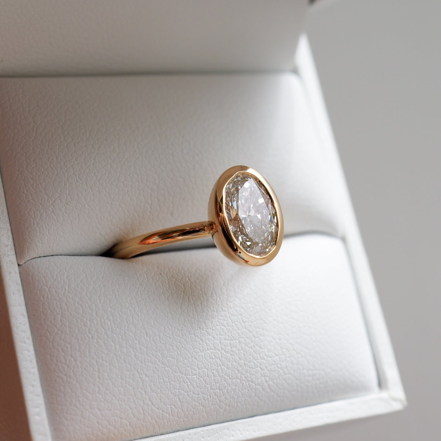 Gold ring with an oval diamond in a bezel setting, displayed in an open white jewellery box. Ideal for luxury engagement rings.